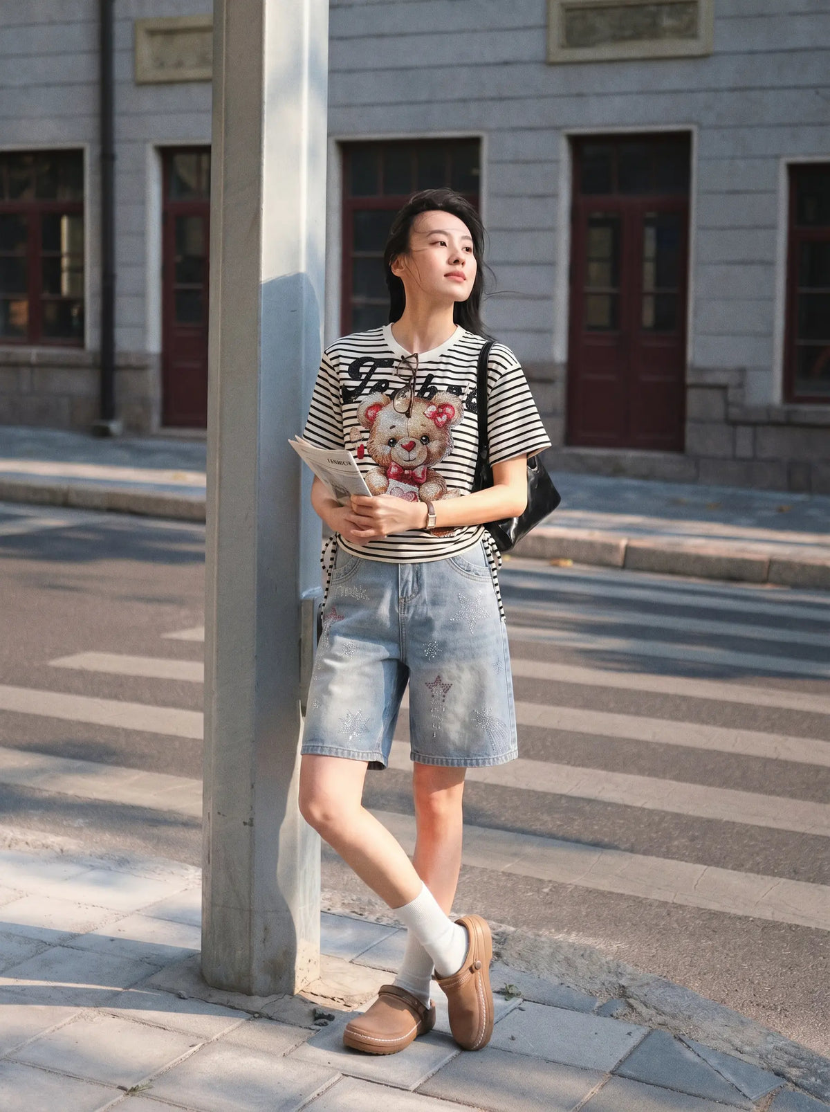 A woman stands on a city sidewalk next to a pole, wearing a black and white striped graphic t-shirt, light-wash denim shorts, white socks, and brown loafers. She is holding a magazine and has a black shoulder bag. TCH Rhinestone Teddy Bear Striped Tie-Side T-Shirt