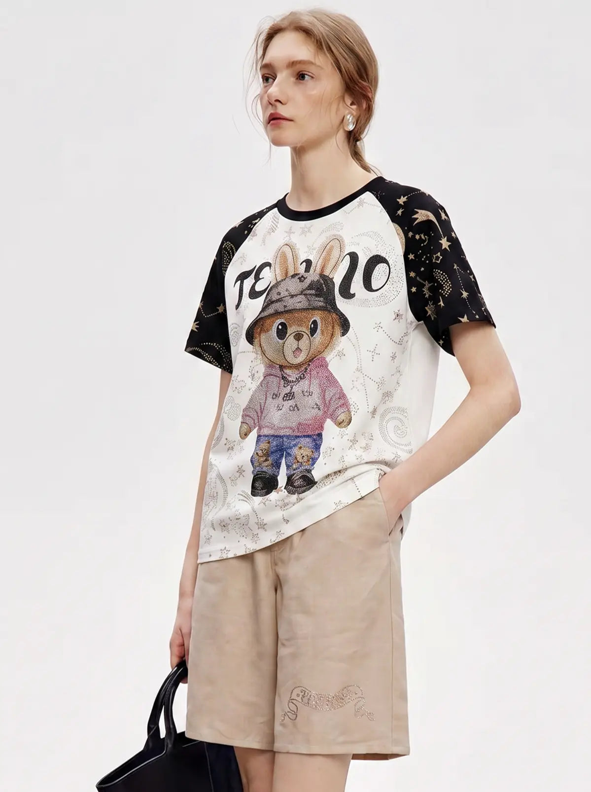 A model wearing a short-sleeve graphic t-shirt with black and off-white panels, featuring a cartoon rabbit illustration and the text 'TELL ME', paired with beige shorts and holding a black handbag against a light gray background. TCH Rhinestone Teddy Bear Techno Raglan T-Shirt