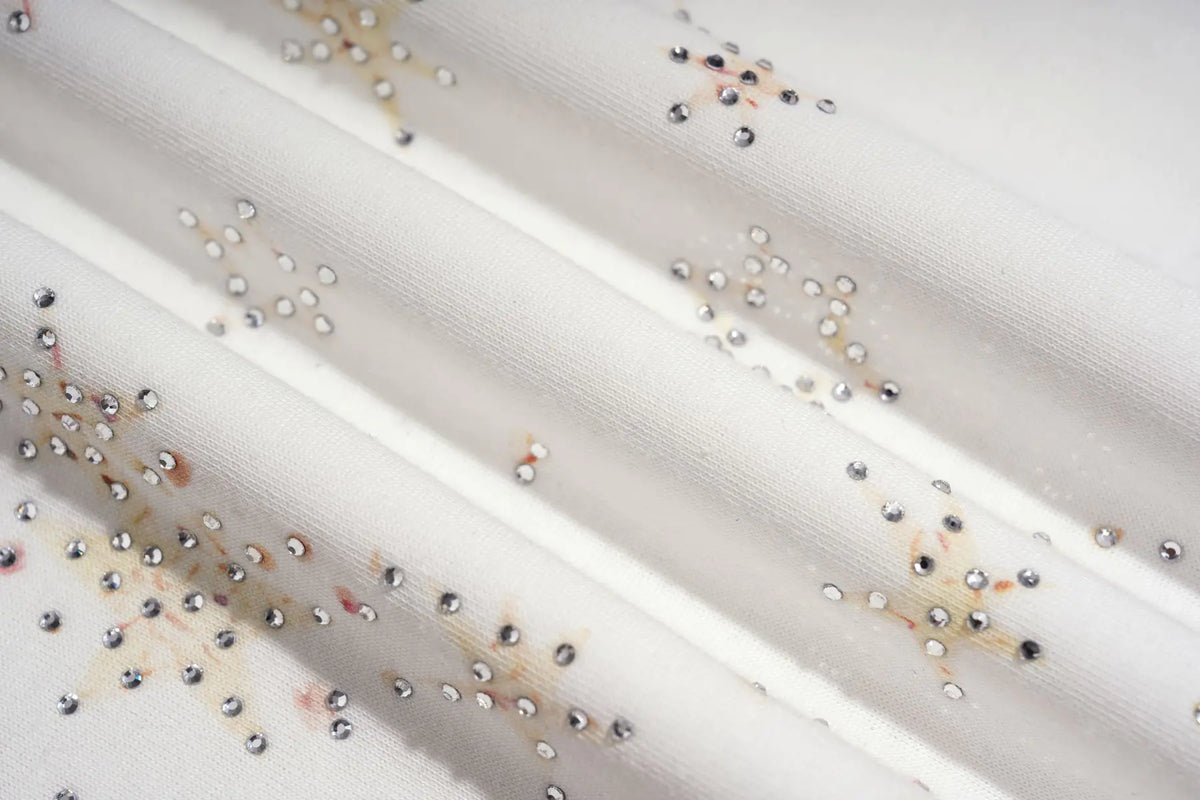 A close-up of a white or off-white sheer fabric with scattered small metallic rhinestones and a few faint brownish marks. TCH Rhinestone Teddy Bear Techno Raglan T-Shirt