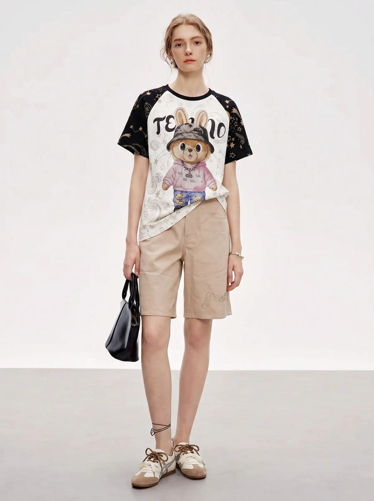 A model stands against a light gray studio background wearing a graphic T-shirt with black lace-embroidered sleeves, beige shorts, beige shoes, and carrying a black handbag. The T-shirt features a white body with a cartoon rabbit graphic and the text 'TEBONO'. TCH Rhinestone Teddy Bear Techno Raglan T-Shirt