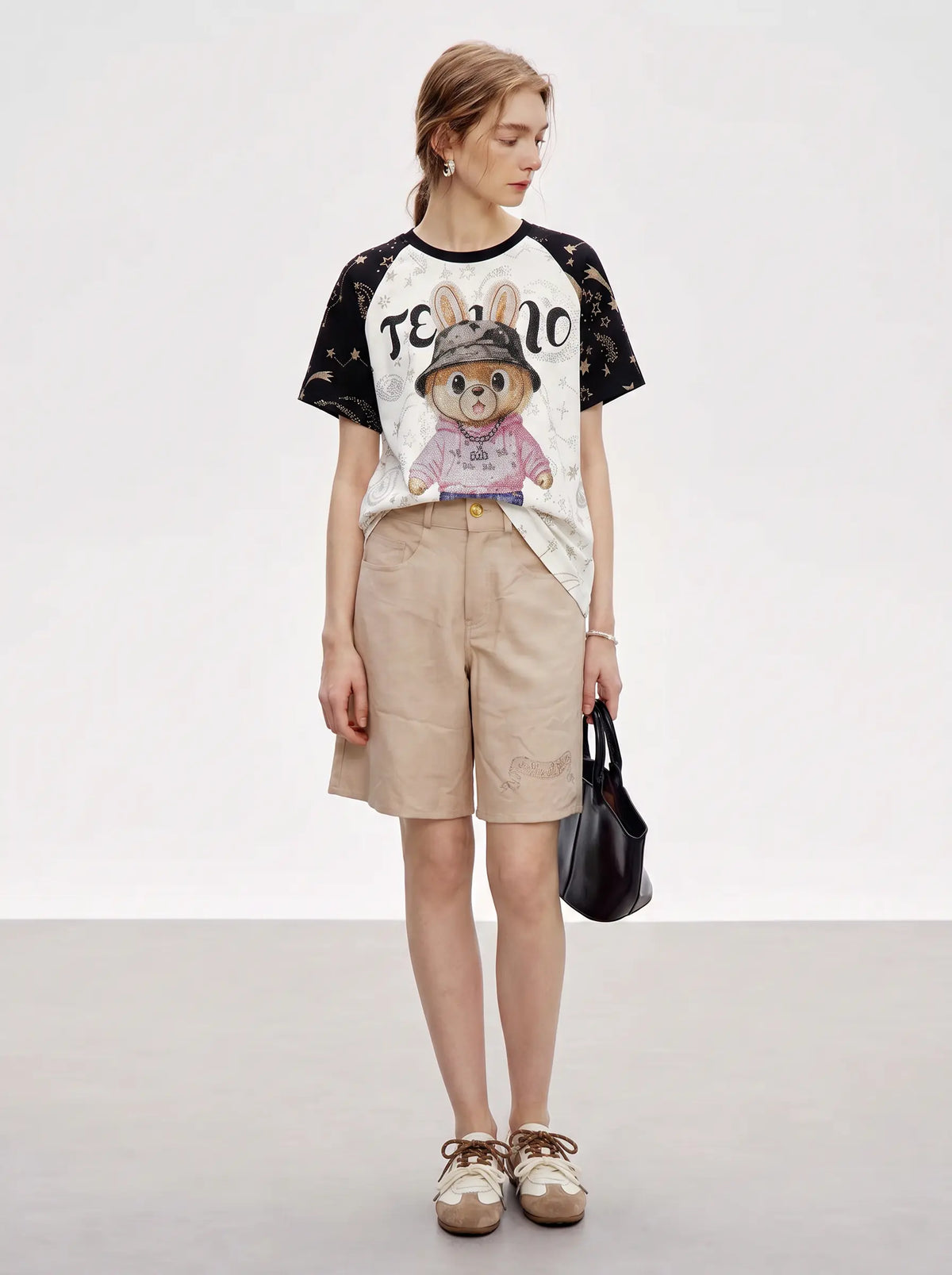 A woman stands in a studio wearing a graphic T-shirt with black sleeves and a white body featuring a cartoon rabbit design and the word 'TEEN', paired with beige shorts, beige shoes, and a black handbag. TCH Rhinestone Teddy Bear Techno Raglan T-Shirt