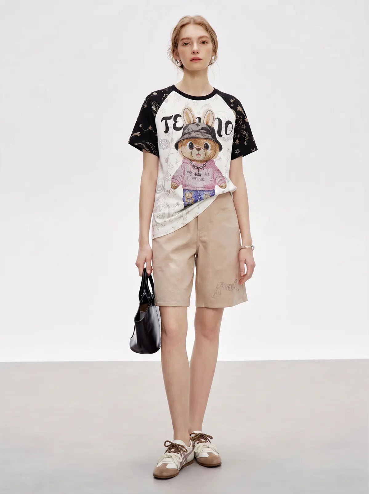 A model stands against a light gray studio background wearing a graphic T-shirt with black sleeves and a white body featuring a cartoon rabbit design and the text 'TEENAGE', paired with beige shorts, brown shoes, and holding a black handbag. TCH Rhinestone Teddy Bear Techno Raglan T-Shirt
