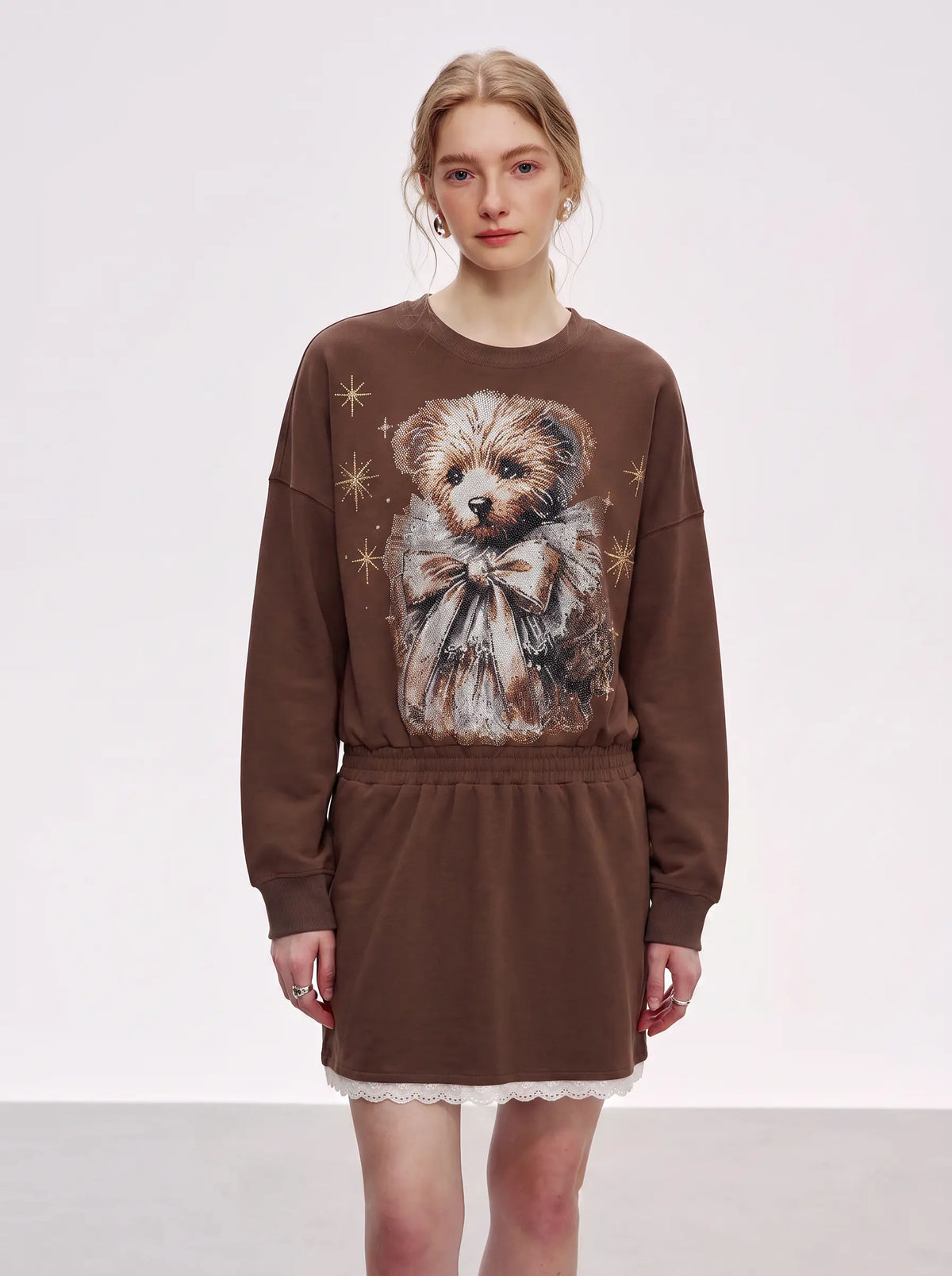 A model wearing a brown crewneck sweatshirt dress featuring a large graphic print of a small dog wearing a bow tie and surrounded by gold stars, with a white lace hem. TCH Rhinestone Teddy Bear Techno Sweatshirt Dress