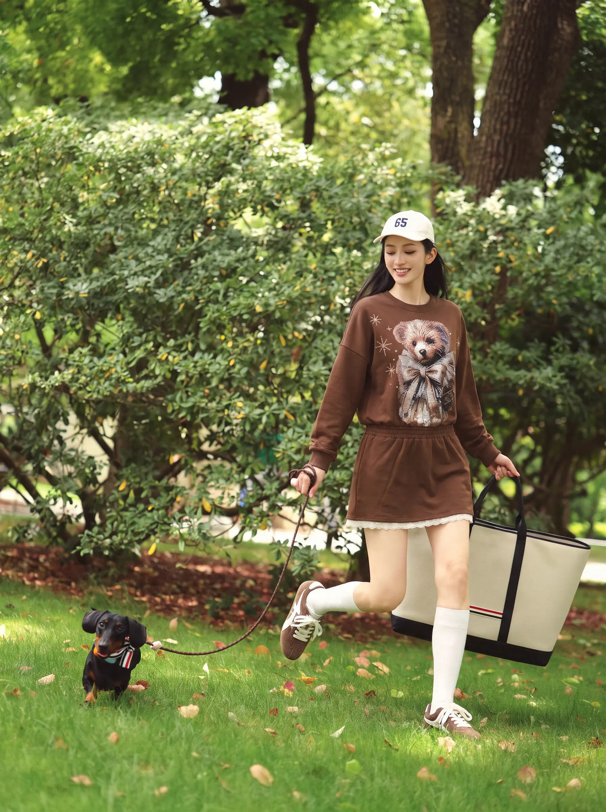 A woman walking in a park with a small black dog on a leash, wearing a brown long-sleeve sweatshirt with a dog graphic, a matching brown mini skirt, white knee-high socks, and carrying a large beige tote bag. TCH Rhinestone Teddy Bear Techno Sweatshirt Dress