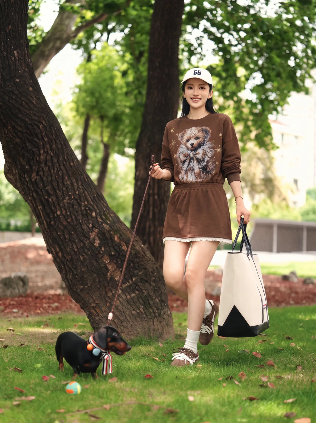 A woman walks on a grassy park path with a small black dog, wearing a brown long-sleeve dress with a bear graphic on the front, a white cap, and carrying a white and black tote bag. TCH Rhinestone Teddy Bear Techno Sweatshirt Dress