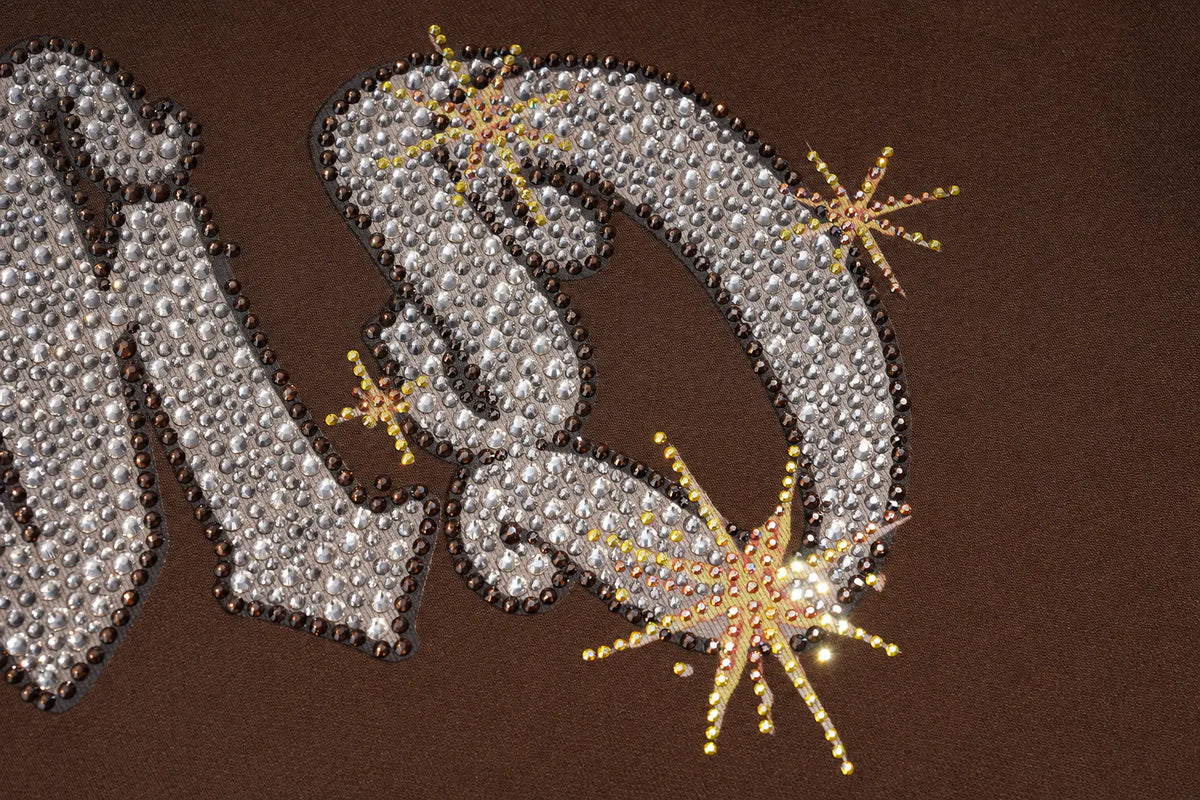 A close-up of a brown garment featuring a large, stylized silver letter 'M' embellished with clear rhinestones, accented by gold-toned starburst rhinestone designs. TCH Rhinestone Teddy Bear Techno Sweatshirt Dress