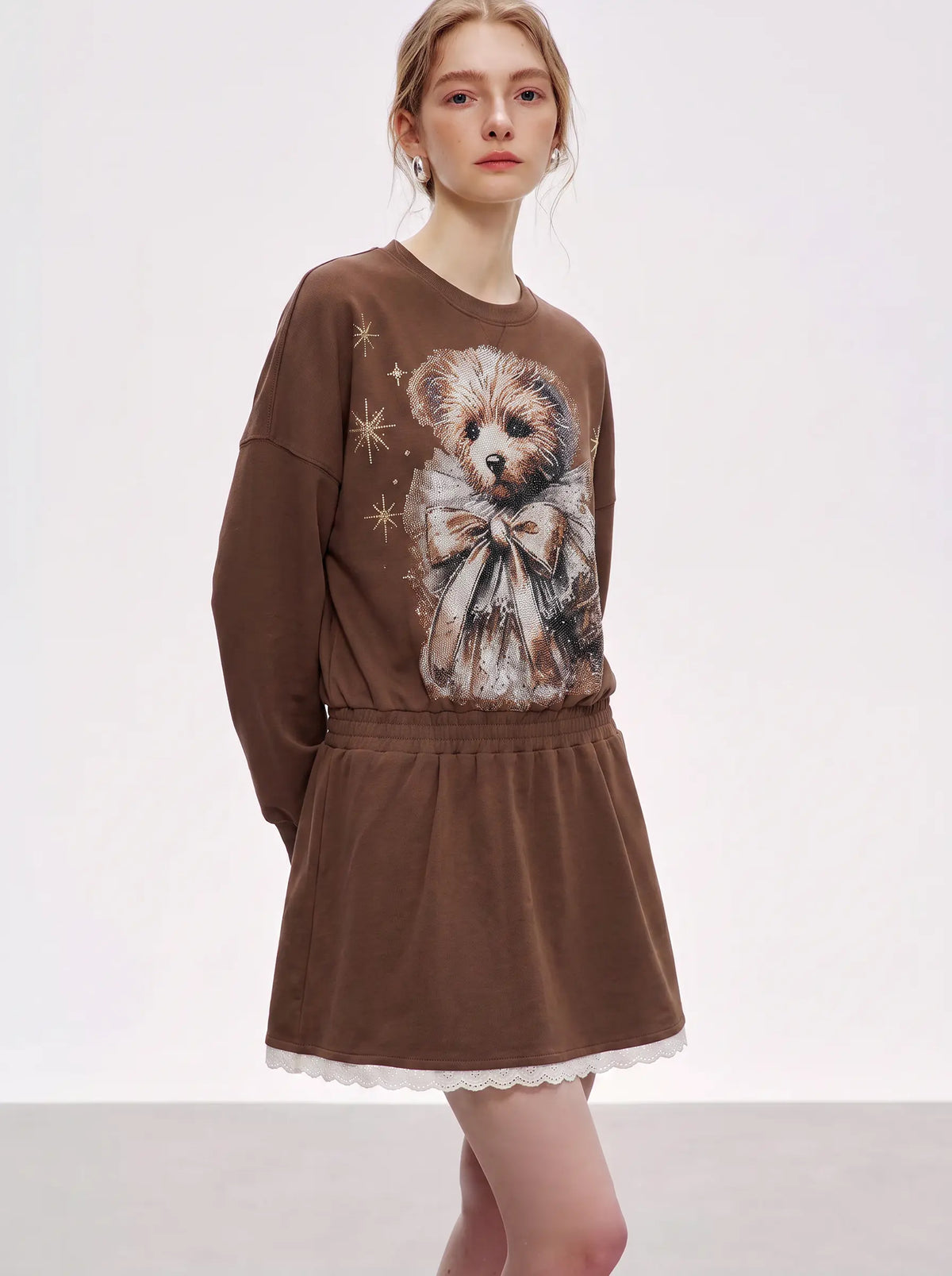 A model wearing a brown long-sleeve dress featuring a large teddy bear graphic on the front, a gathered elastic waist, and a white scalloped hem TCH Rhinestone Teddy Bear Techno Sweatshirt Dress