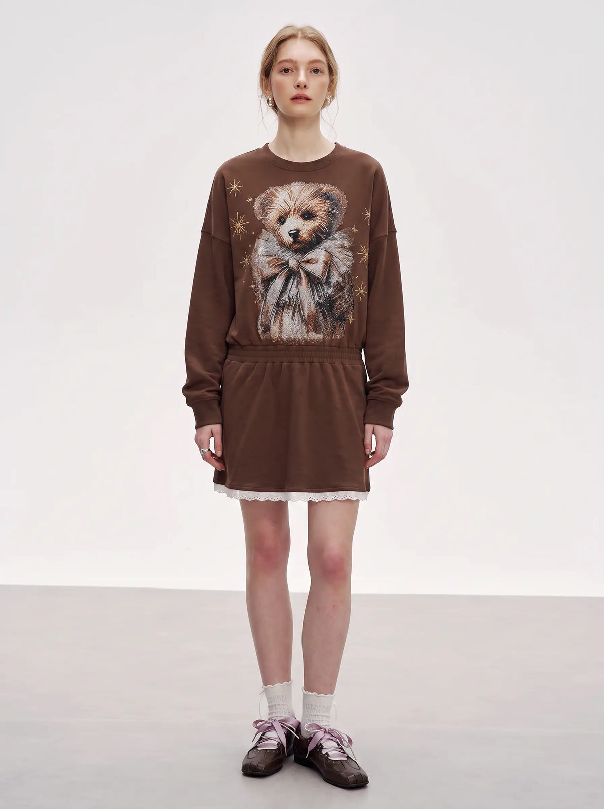 A model stands against a plain light gray studio background wearing a brown long-sleeve sweatshirt dress with a large teddy bear graphic on the front, white lace trim at the hem, white socks, and brown lace-up shoes. TCH Rhinestone Teddy Bear Techno Sweatshirt Dress