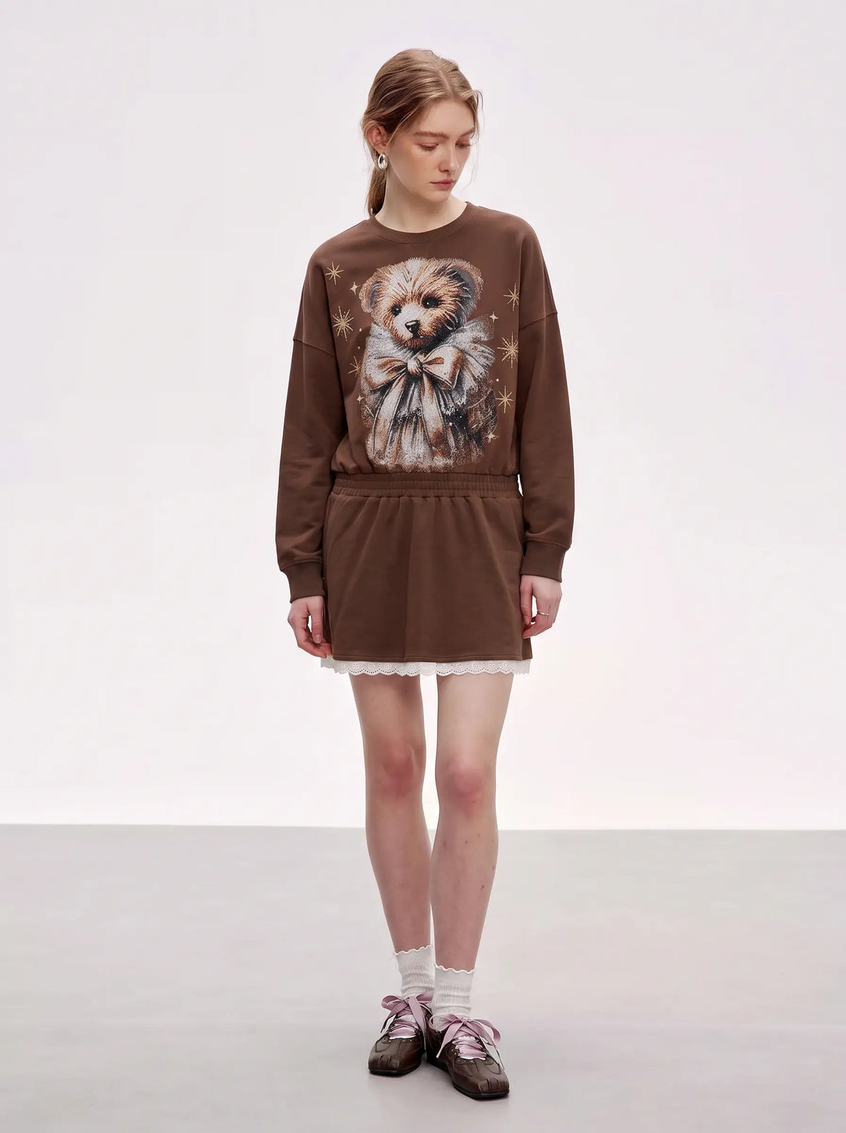 A model stands in a studio wearing a brown sweatshirt dress with a large graphic of a fluffy dog on the front, paired with white socks and brown shoes. TCH Rhinestone Teddy Bear Techno Sweatshirt Dress