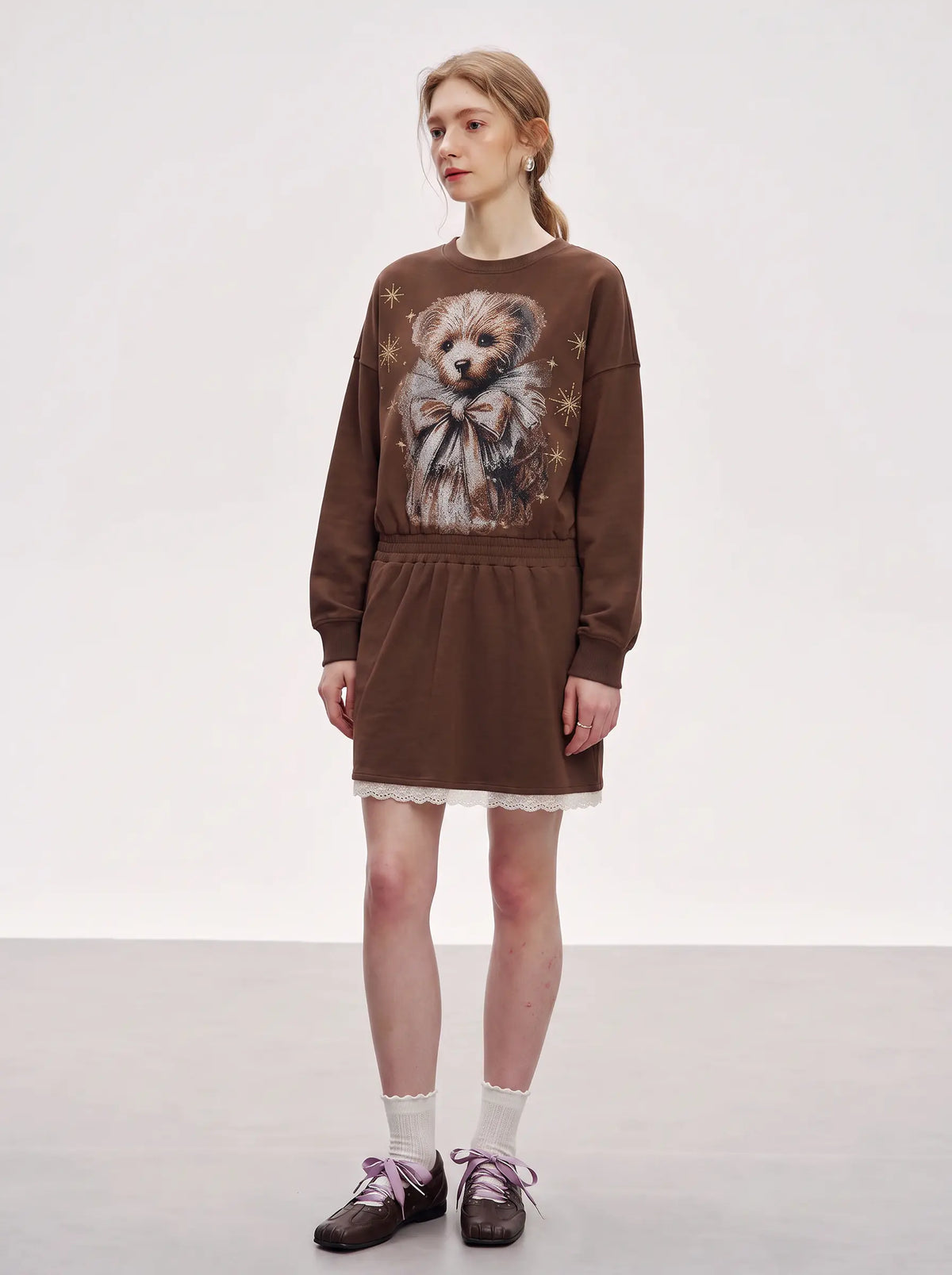 A model stands in a studio wearing a brown long-sleeve sweatshirt dress with a large teddy bear graphic on the front, white lace trim at the hem, white socks, and dark brown shoes. TCH Rhinestone Teddy Bear Techno Sweatshirt Dress