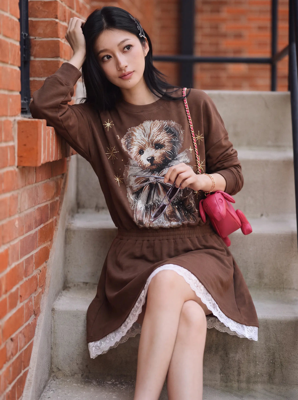 A woman sits on outdoor concrete steps next to a red brick wall, wearing a brown long-sleeve dress with a large dog graphic on the front, white lace trim at the hem, and holding a pink handbag. TCH Rhinestone Teddy Bear Techno Sweatshirt Dress