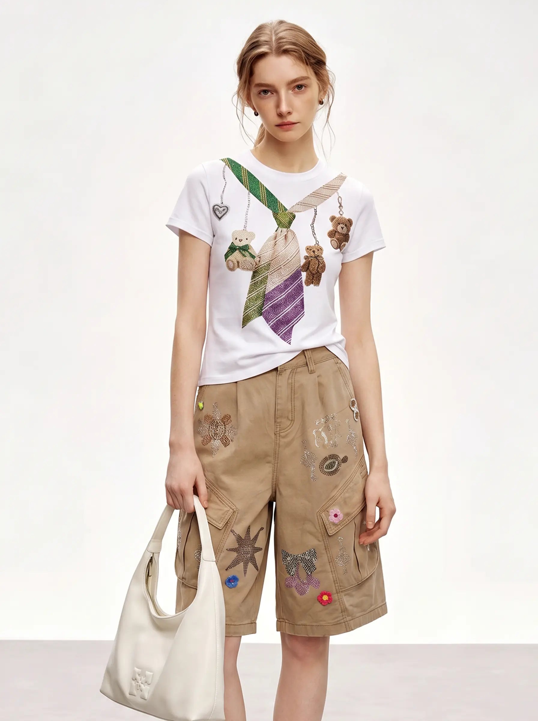 A model wearing a white graphic t-shirt with teddy bear keychains and a purple striped tie print, paired with beige cargo shorts featuring colorful embroidered patches, holding a cream-colored leather shoulder bag against a plain white studio background. TCH Rhinestone Teddy Bear Tie Graphic White T-Shirt