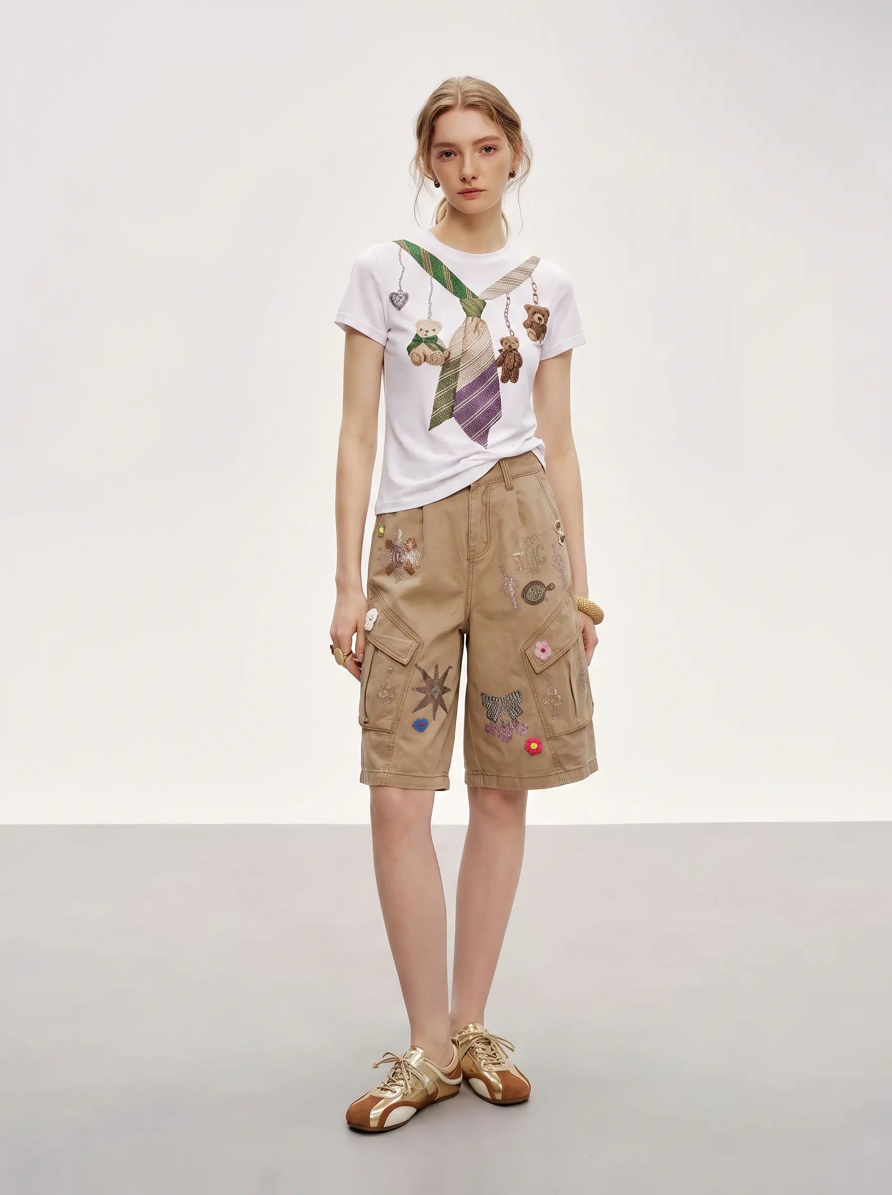 A model stands against a light gray studio background wearing a white graphic T-shirt, tan cargo shorts with colorful embroidered patches, and metallic gold sandals. TCH Rhinestone Teddy Bear Tie Graphic White T-Shirt