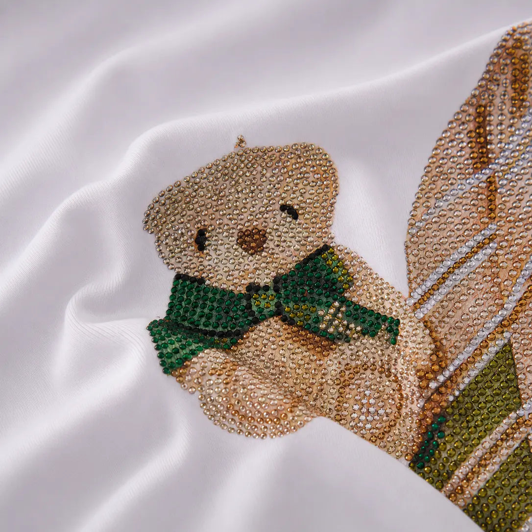 A close-up of a white satin fabric featuring an embroidered teddy bear motif. The bear is made of beige beads and wears a green beaded scarf, with a plaid pattern in beige, green, and white visible on the right side. TCH Rhinestone Teddy Bear Tie Graphic White T-Shirt