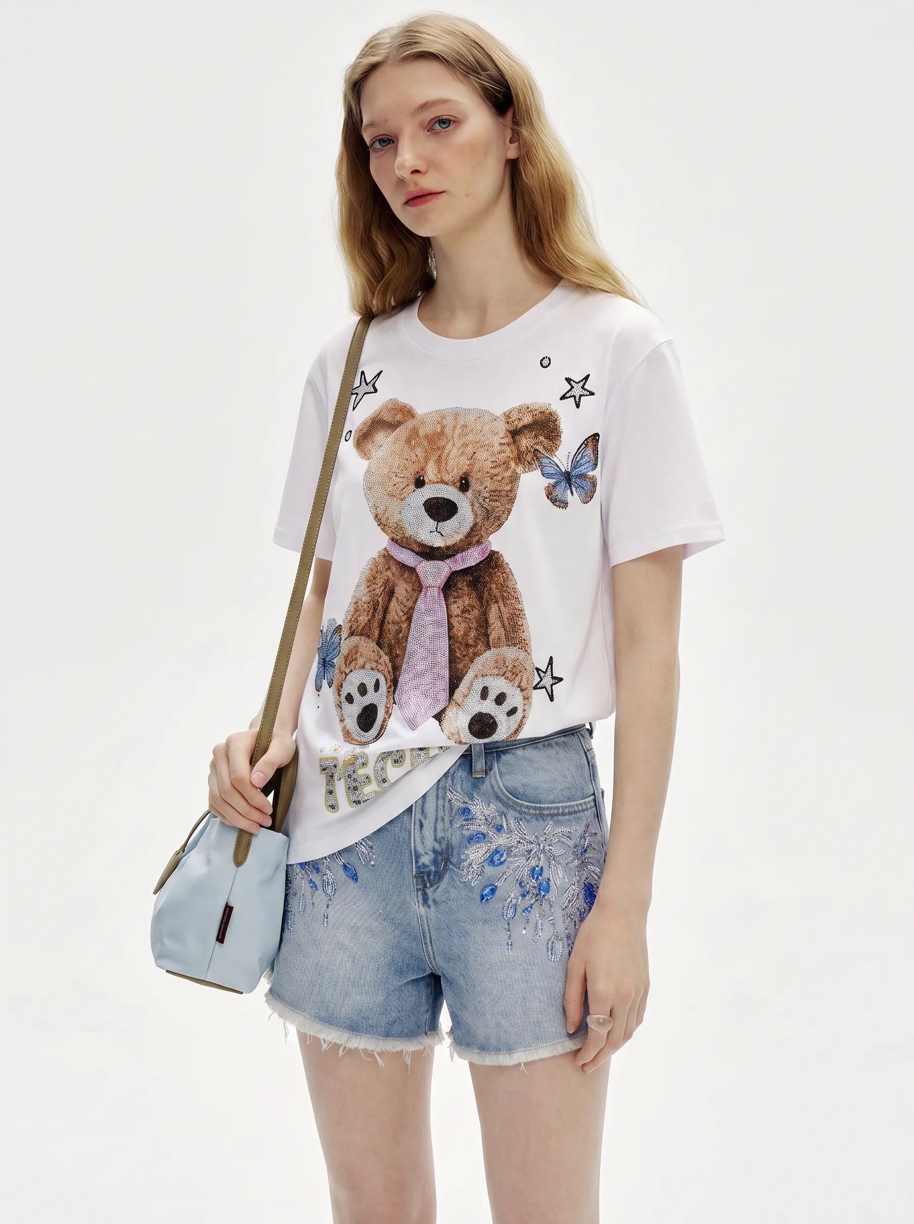 A model wearing a white short-sleeve t-shirt with a large brown teddy bear graphic, paired with light blue denim shorts and carrying a light blue shoulder bag against a plain white background. TCH Rhinestone Teddy Bear White T-Shirt