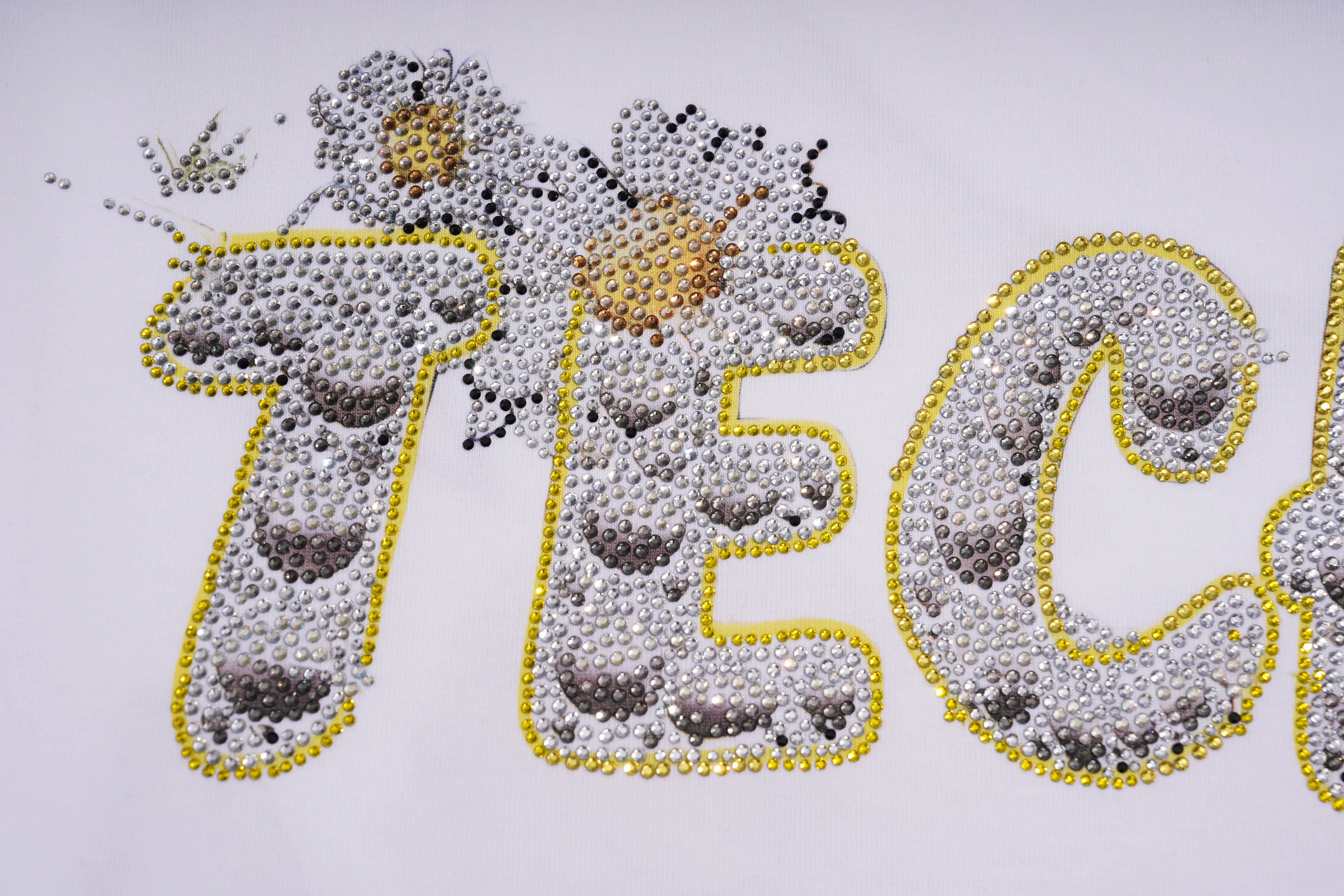 A close-up of large decorative letters outlined in yellow and filled with white, gray, and black rhinestones, with a small gold flower detail above, on a plain light background. TCH Rhinestone Teddy Bear White T-Shirt