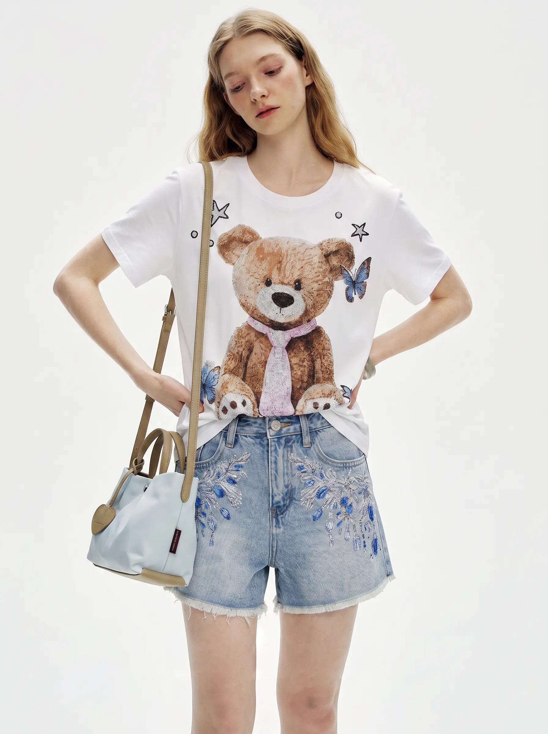 A model wearing a white graphic t-shirt with a large teddy bear print, light-wash denim shorts with blue floral embroidery, and carrying a light blue shoulder bag against a plain white background. TCH Rhinestone Teddy Bear White T-Shirt