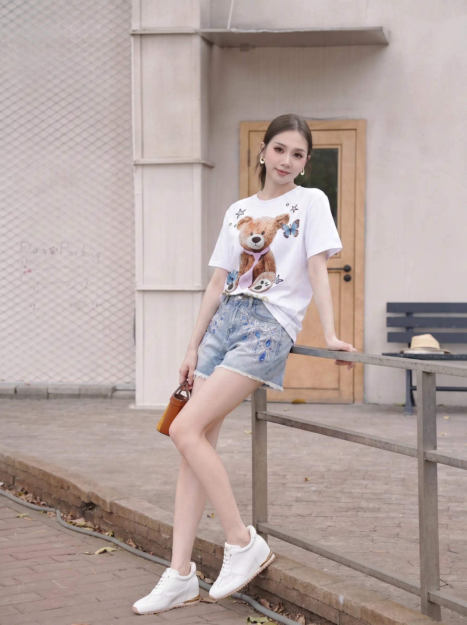 A woman posing outdoors in a white graphic t-shirt with a teddy bear print, light blue denim shorts, and white sneakers, leaning on a metal railing. TCH Rhinestone Teddy Bear White T-Shirt