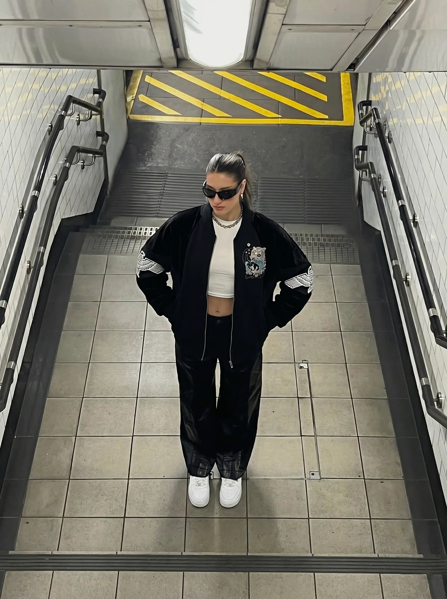 A person standing in a subway station wearing a black zip-up bomber jacket with white sleeve stripes, black pants, a white crop top, and white sneakers. TCH Rhinestone Teddy Bear Wing Sleeve Bomber Jacket