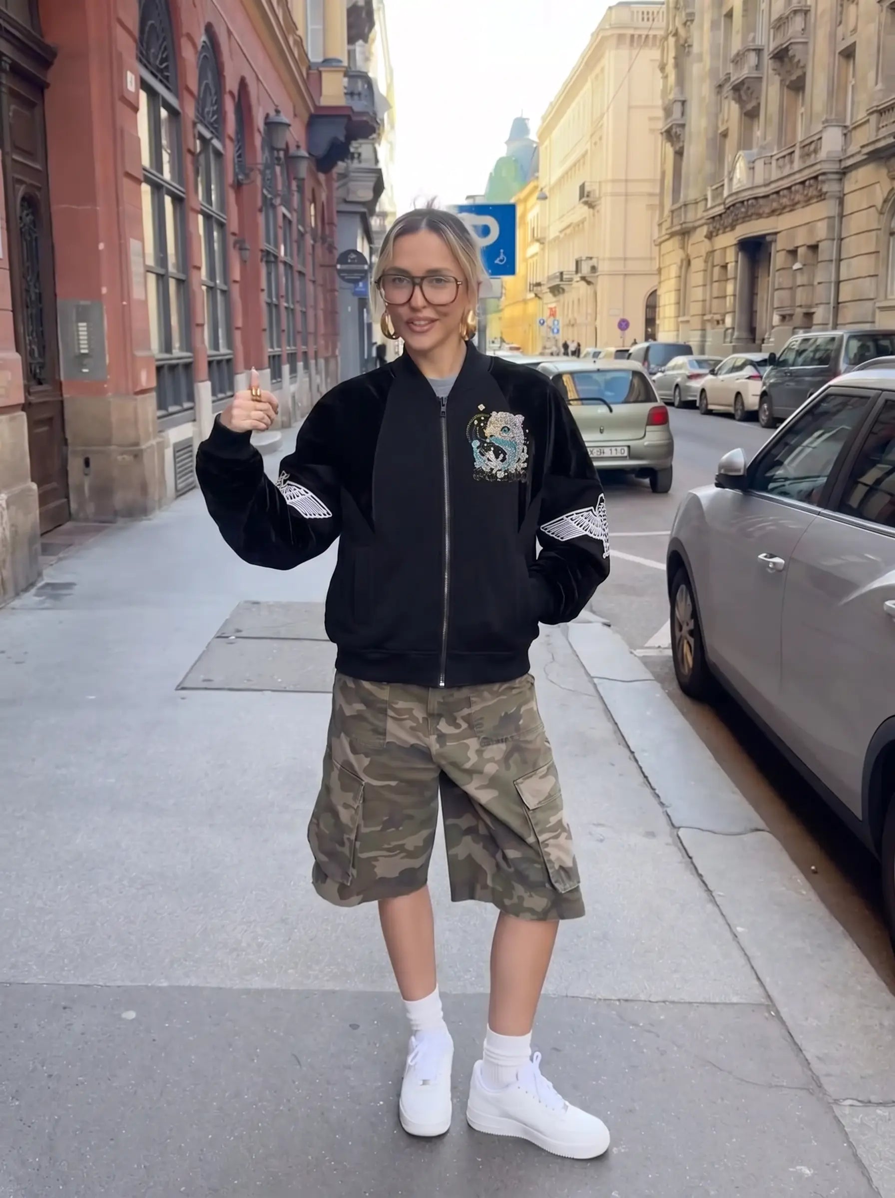 A woman stands on a city sidewalk wearing a black bomber jacket with white sleeve accents, camouflage cargo shorts, white socks, and white sneakers. The background shows urban buildings and parked cars. TCH Rhinestone Teddy Bear Wing Sleeve Bomber Jacket