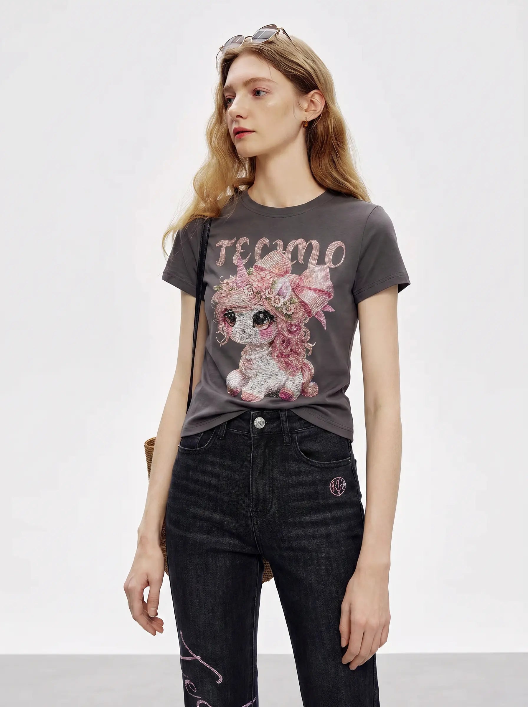 A woman modeling a dark gray graphic t-shirt with a pink cartoon character and the word 'TECMO' printed on the front, paired with dark wash jeans against a plain light gray studio background. TCH Rhinestone Unicorn Graphic Charcoal T-Shirt