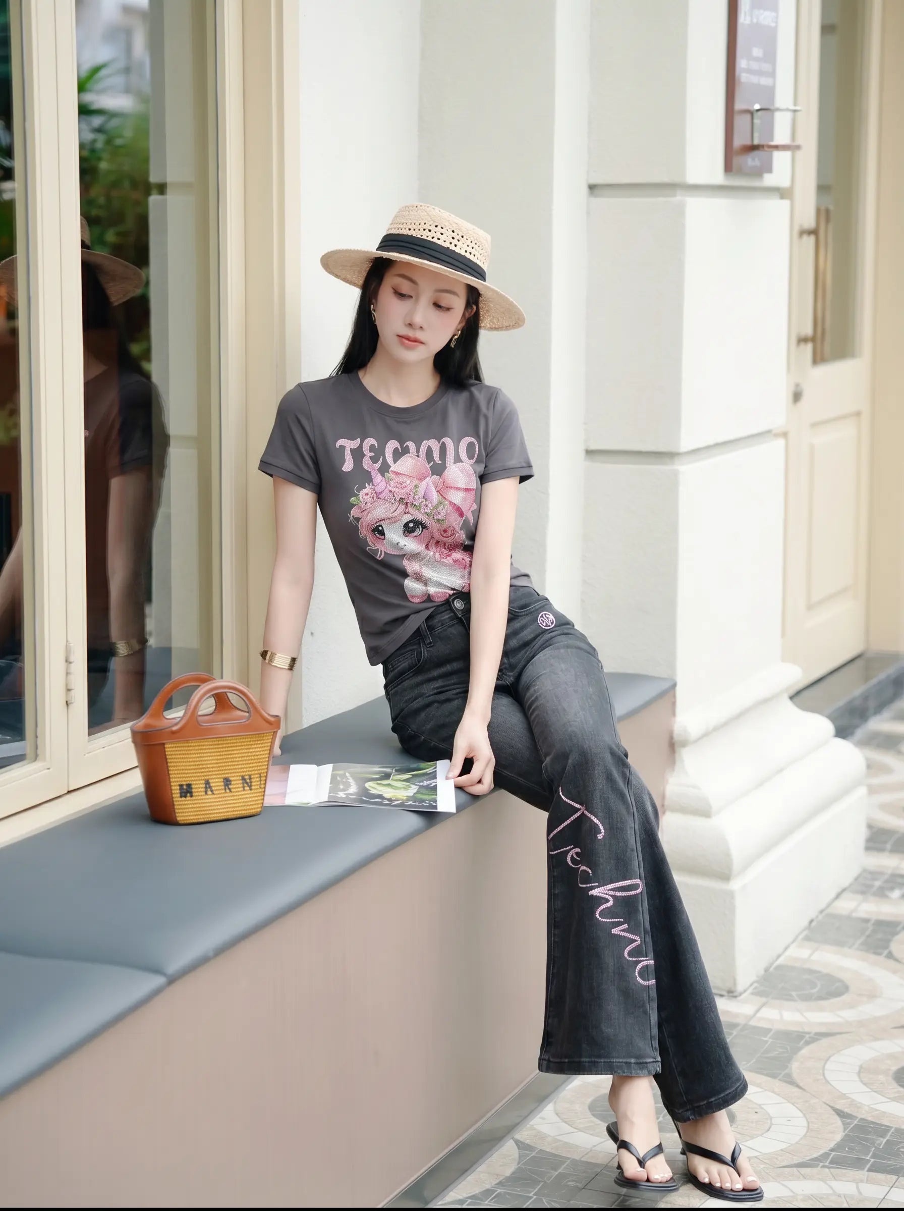 A woman wearing a dark gray graphic t-shirt with a pink graphic and the word 'TECHNO', paired with dark gray wide-leg jeans featuring 'Techno' script, a straw hat, and black sandals, sitting on a beige bench outside a building. TCH Rhinestone Unicorn Graphic Charcoal T-Shirt