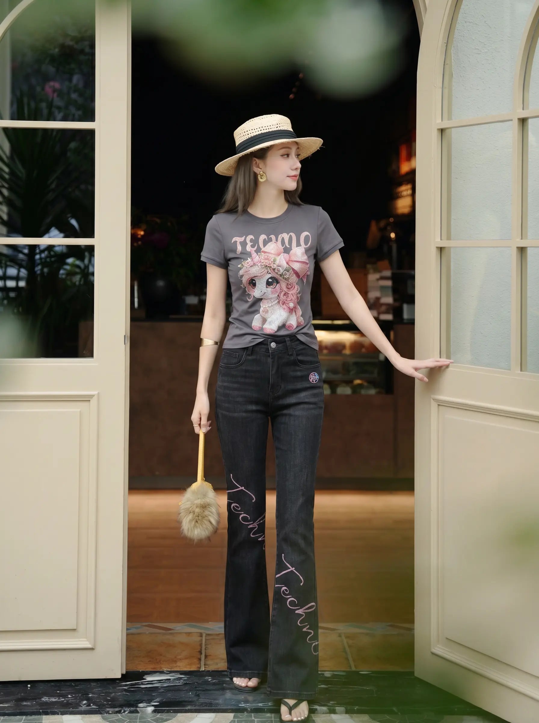 A woman stands in a doorway wearing a dark gray graphic T-shirt with a cartoon bear and the word 'TECMO', paired with dark gray flare jeans, a straw hat, and holding a fluffy beige accessory. TCH Rhinestone Unicorn Graphic Charcoal T-Shirt