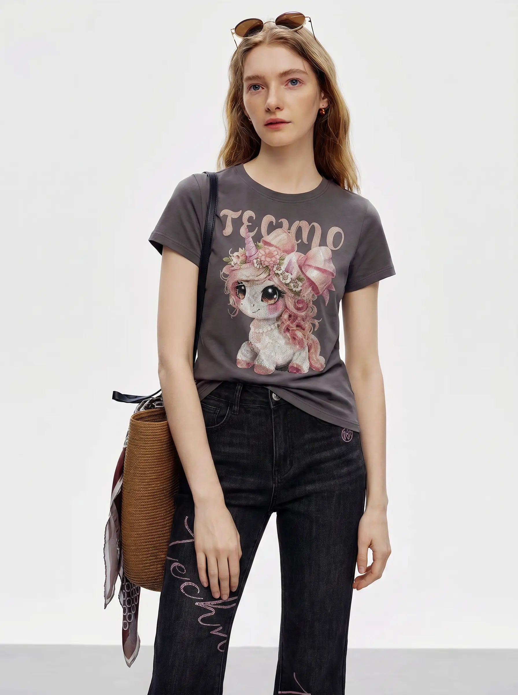 A woman modeling a dark gray graphic t-shirt with a pink and white cartoon bunny and the word 'TEEMO' printed on the front, paired with dark jeans and carrying a brown woven shoulder bag. TCH Rhinestone Unicorn Graphic Charcoal T-Shirt