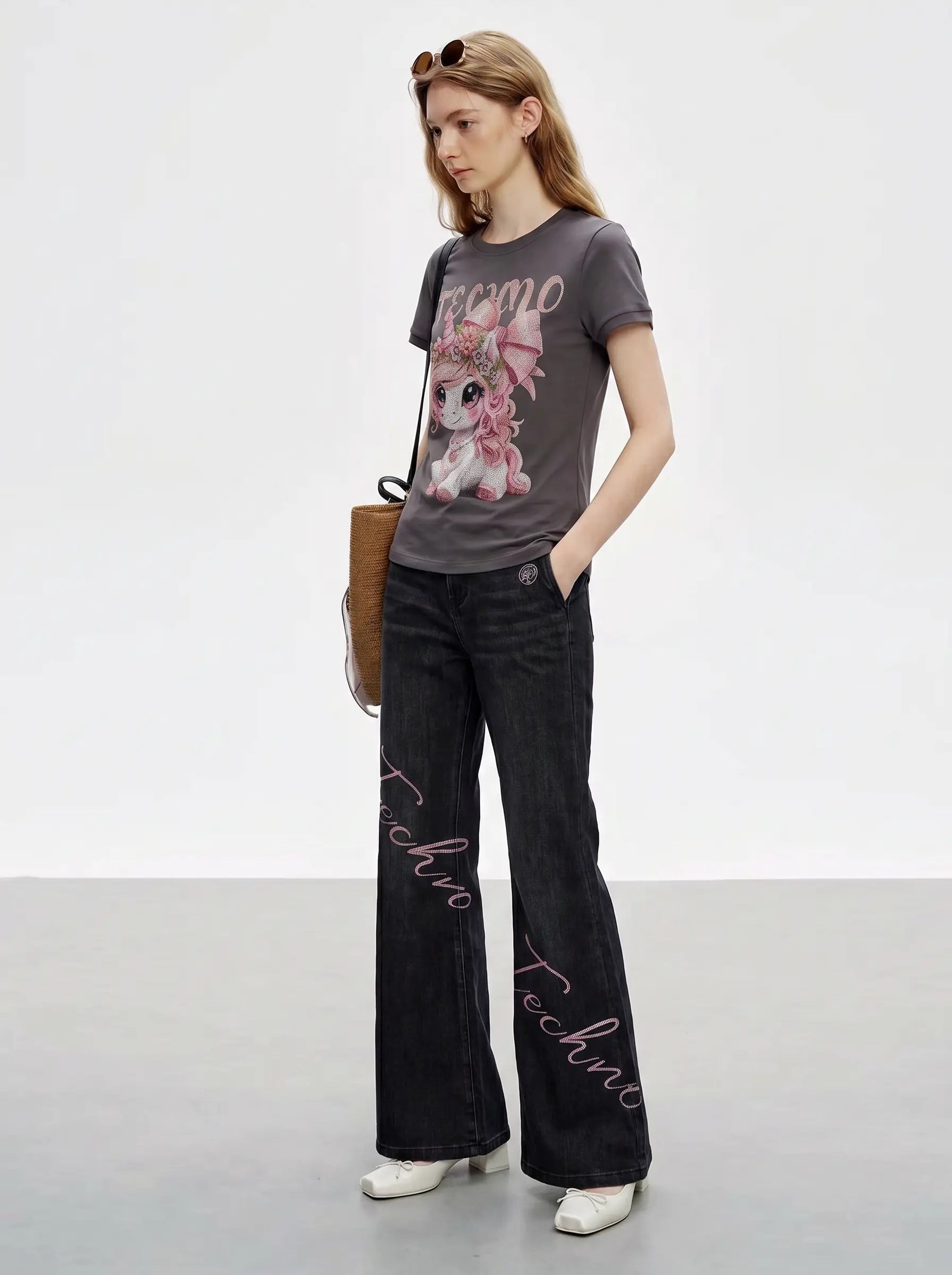 A model stands in a studio wearing a dark gray graphic t-shirt with a pink cartoon character and the word 'REMO', paired with dark gray wide-leg jeans featuring pink script text down the front, white sneakers, and carrying a brown leather bag. TCH Rhinestone Unicorn Graphic Charcoal T-Shirt