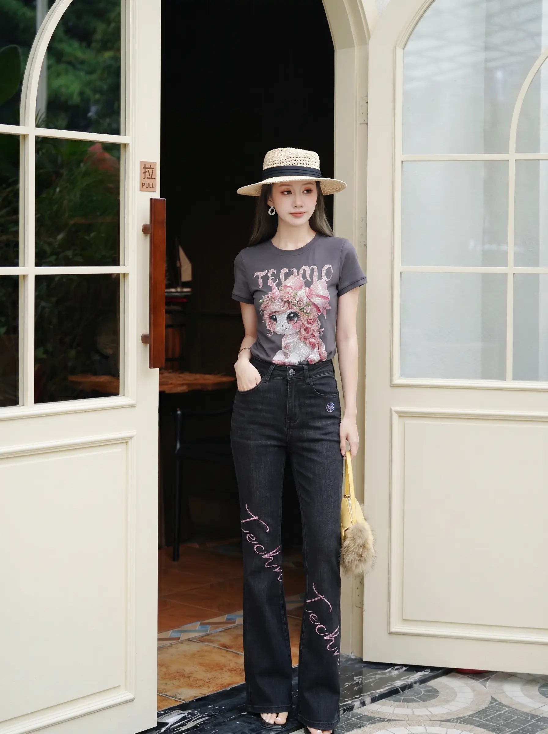 A woman stands in front of open cream-colored arched doors wearing a dark gray graphic T-shirt with a teddy bear print, black wide-leg jeans with script embroidery on the lower legs, a beige straw hat, and carrying a small beige handbag. TCH Rhinestone Unicorn Graphic Charcoal T-Shirt