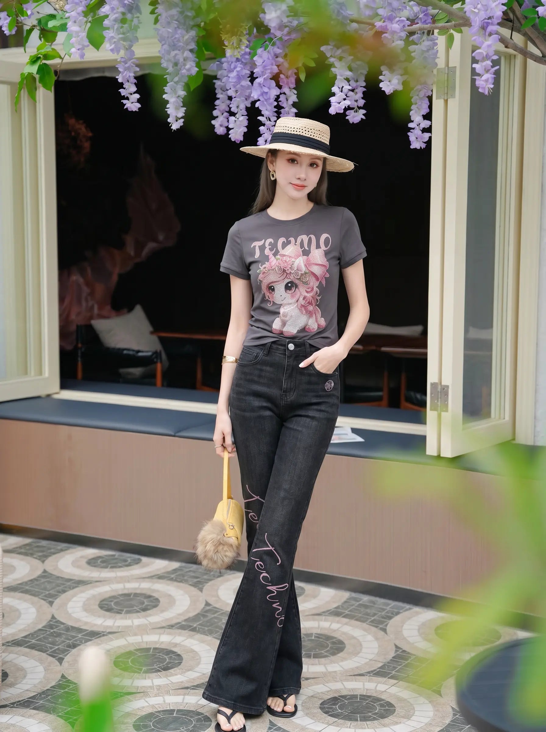 A woman stands outdoors in front of a building entrance wearing a dark gray graphic T-shirt with a pink cat print and the word 'TECMRO' on the chest, paired with dark flare jeans, a straw hat, and carrying a small beige handbag. TCH Rhinestone Unicorn Graphic Charcoal T-Shirt