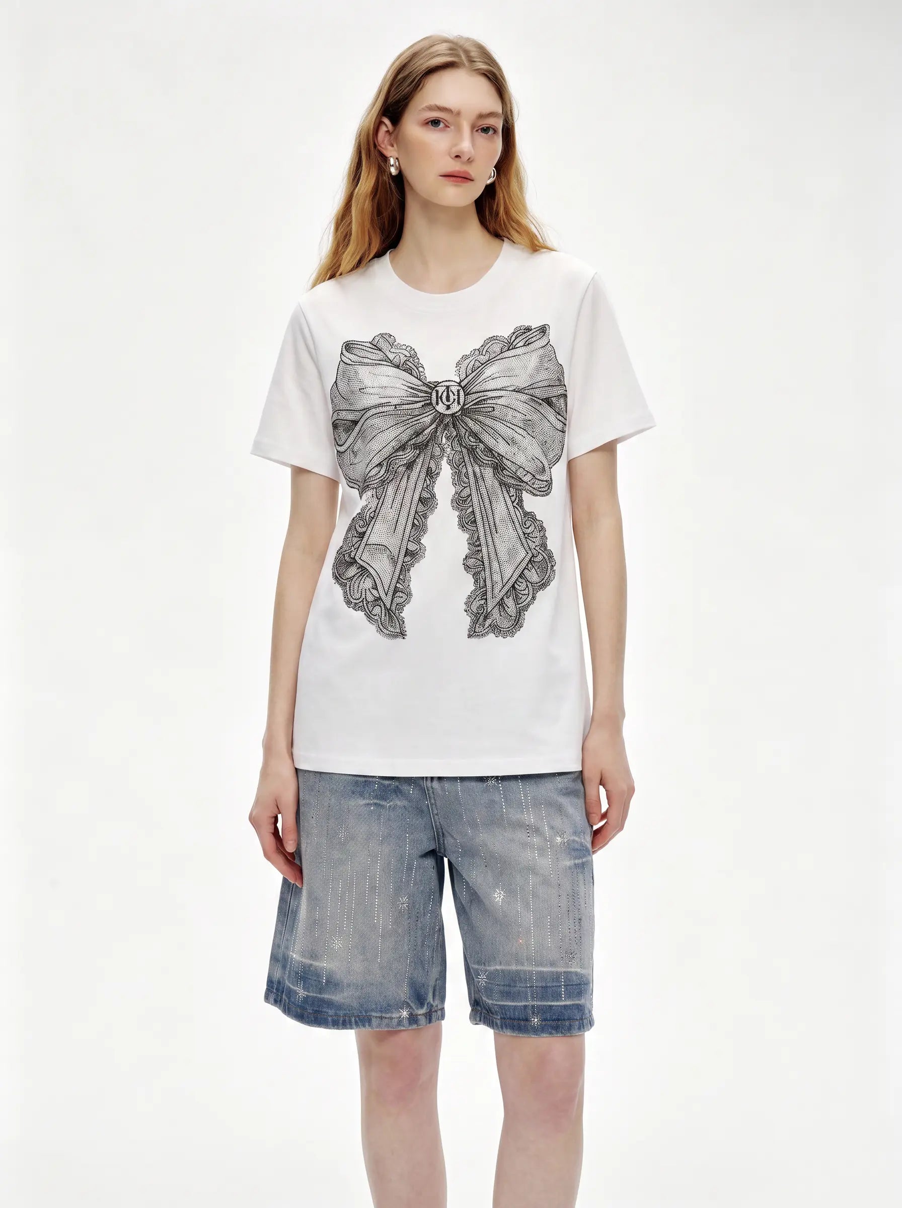 A woman modeling a white short-sleeve T-shirt with a large gray bow graphic on the front, paired with light-wash denim shorts, against a plain white studio background. TCH Rhinestone Vintage Bow Graphic White T-Shirt