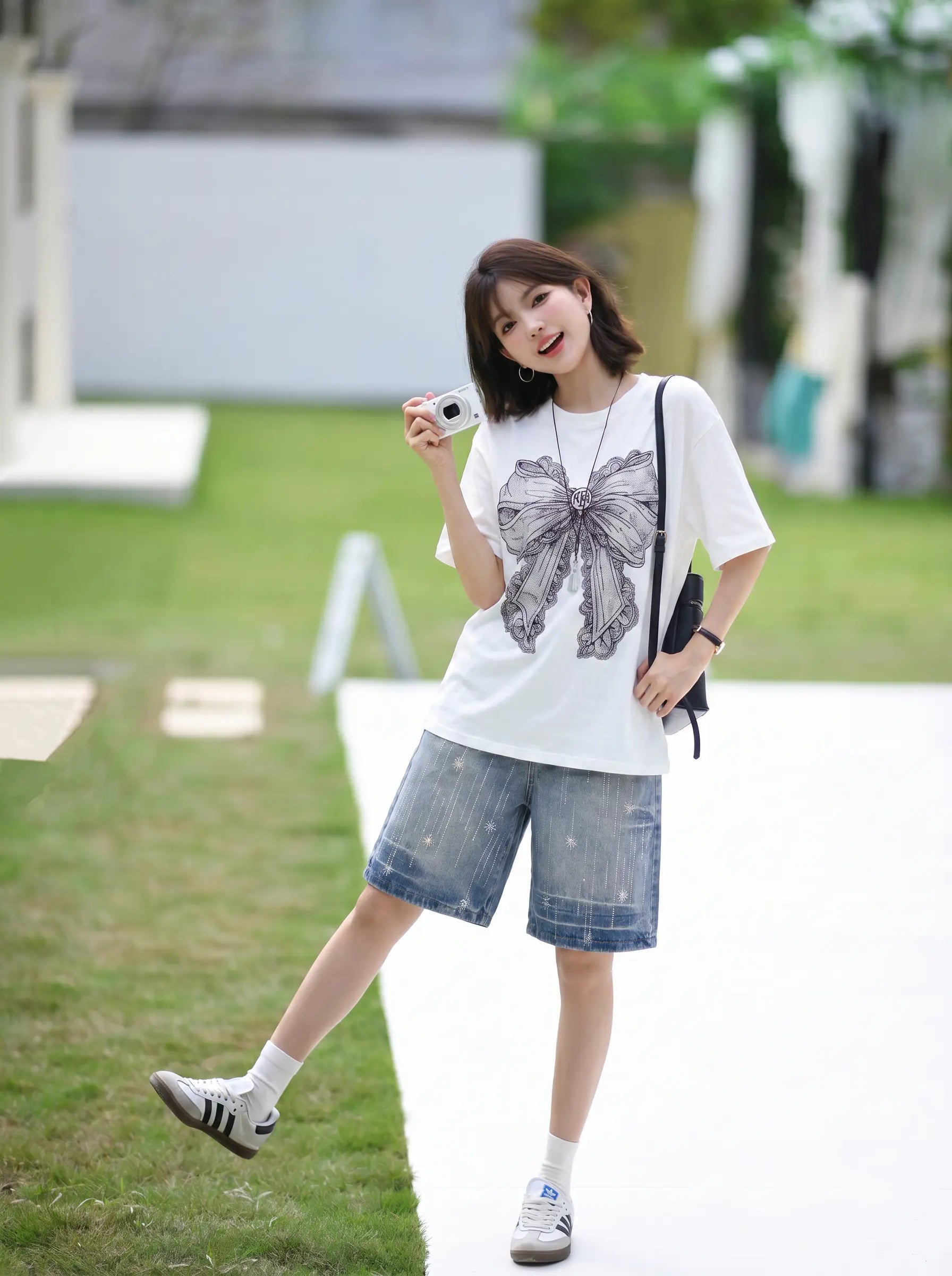 A woman posing outdoors in a white short-sleeve t-shirt with a large gray bow graphic, paired with denim shorts, white socks, and sneakers. TCH Rhinestone Vintage Bow Graphic White T-Shirt