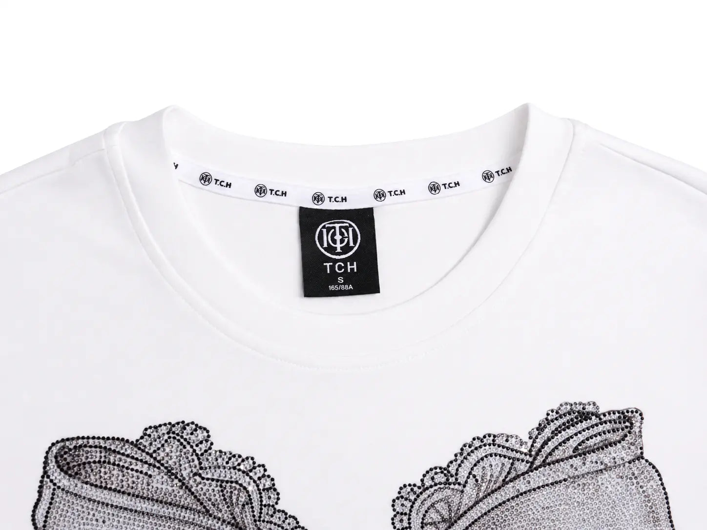 Close-up of a white crewneck T-shirt by TCH, showing the black neck label and the top of a black lace and dot pattern graphic on the front. TCH Rhinestone Vintage Bow Graphic White T-Shirt