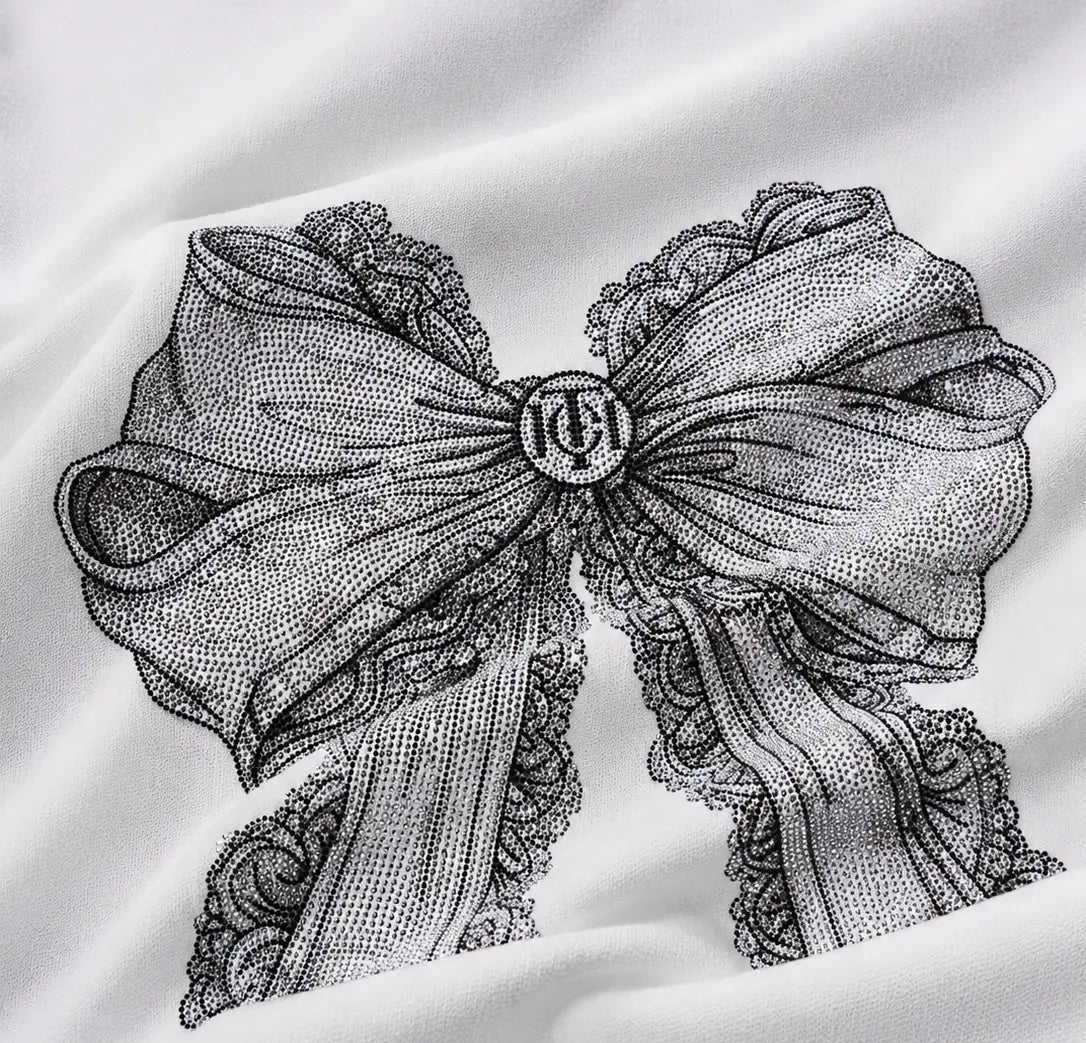 A close-up of a white fabric surface featuring a large black dotted illustration of a ribbon bow with lace edges and a circular monogram at the center. TCH Rhinestone Vintage Bow Graphic White T-Shirt