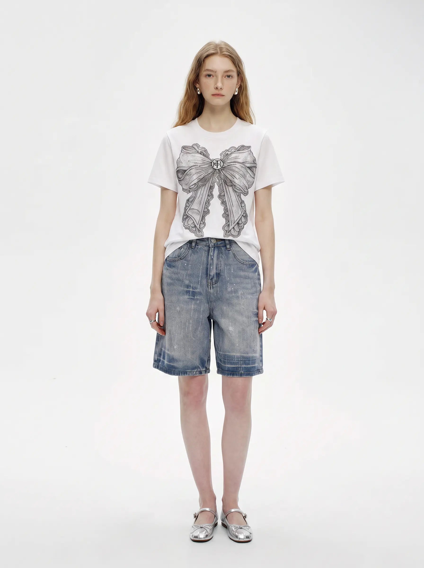 A model stands against a plain white studio background wearing a white short-sleeve T-shirt with a large gray bow graphic on the front, light-wash blue denim shorts, and silver flats. TCH Rhinestone Vintage Bow Graphic White T-Shirt