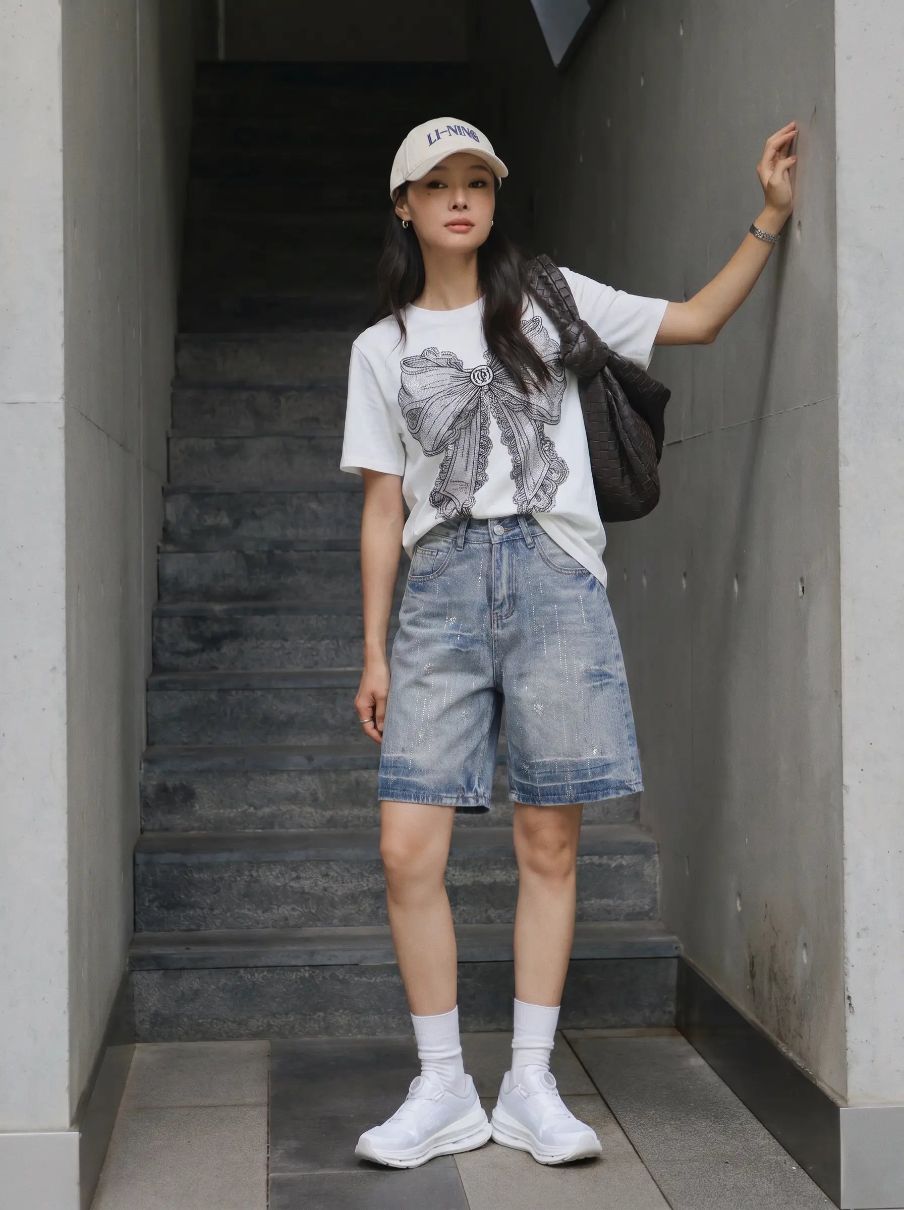 A woman stands in a concrete stairwell wearing a white graphic T-shirt, light-wash denim shorts, white socks, white sneakers, and a white baseball cap, with a dark shoulder bag. TCH Rhinestone Vintage Bow Graphic White T-Shirt