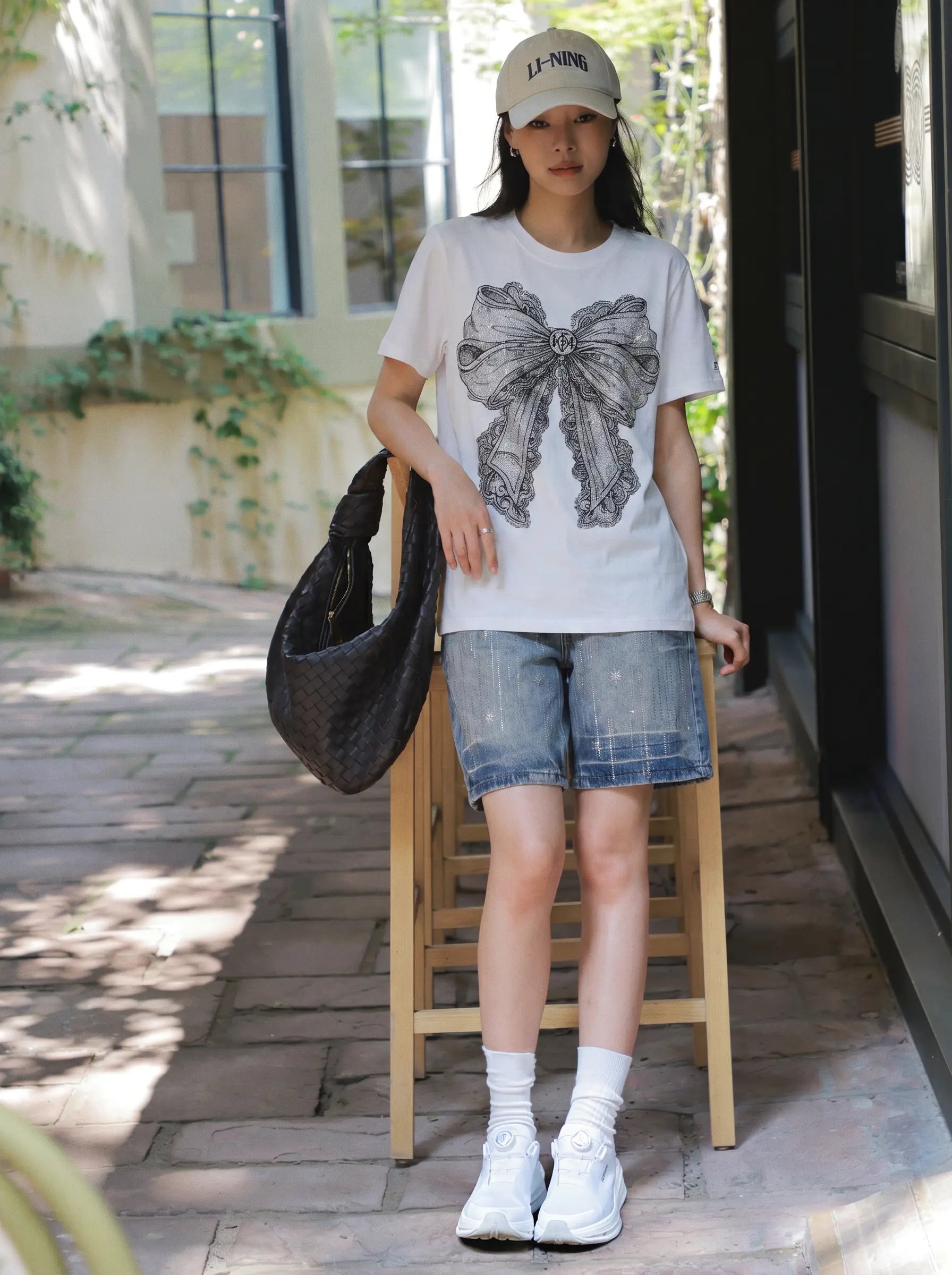 A woman stands outdoors on a stone walkway wearing a white graphic T-shirt with a large gray bow print, denim shorts, white socks, white sneakers, and a beige cap. She is holding a black woven shoulder bag and leaning against a wooden stool. TCH Rhinestone Vintage Bow Graphic White T-Shirt