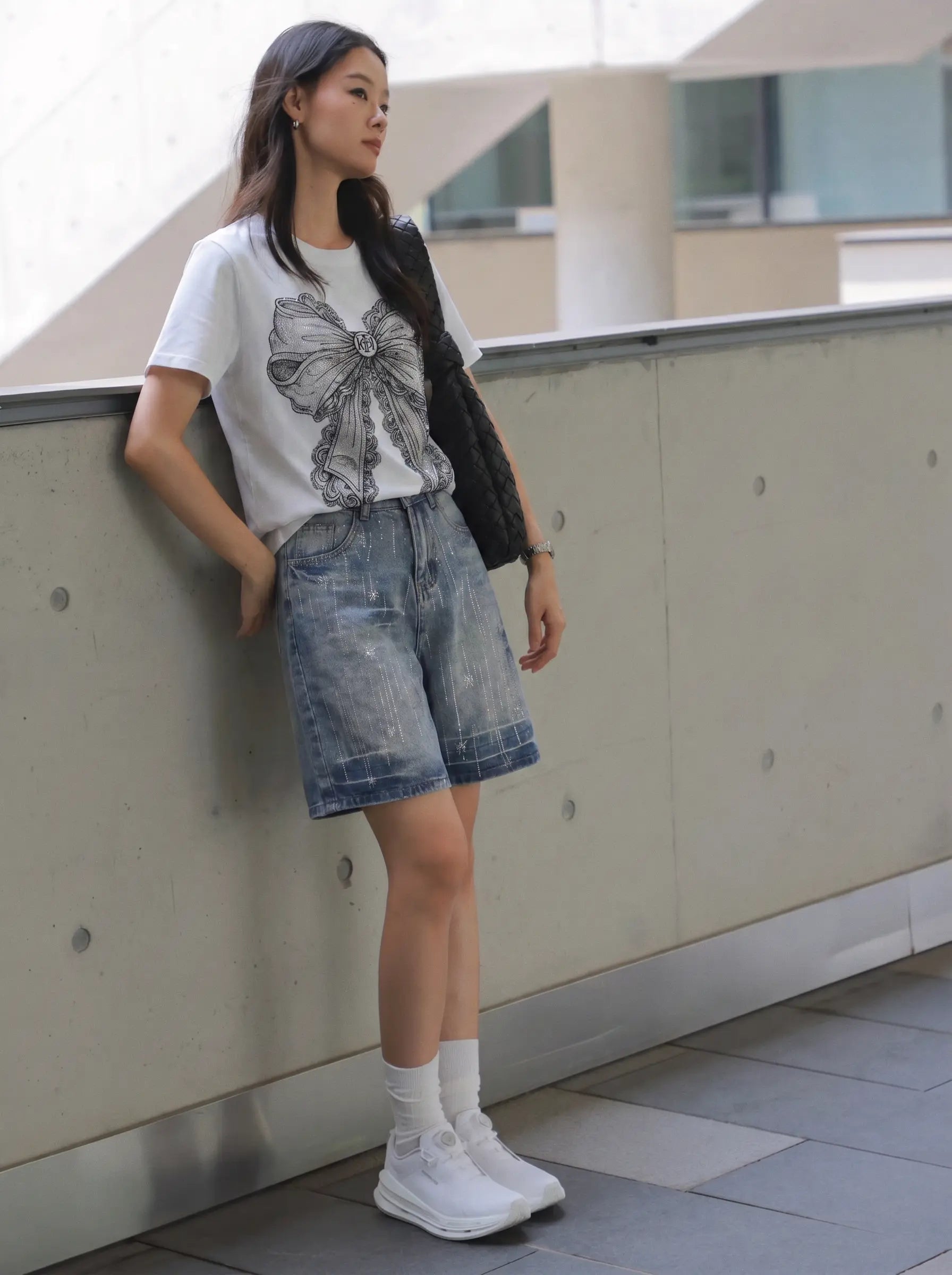 A woman stands outdoors leaning against a concrete wall, wearing a white graphic T-shirt, light-wash denim shorts, white socks, and white sneakers, with a dark shoulder bag. TCH Rhinestone Vintage Bow Graphic White T-Shirt