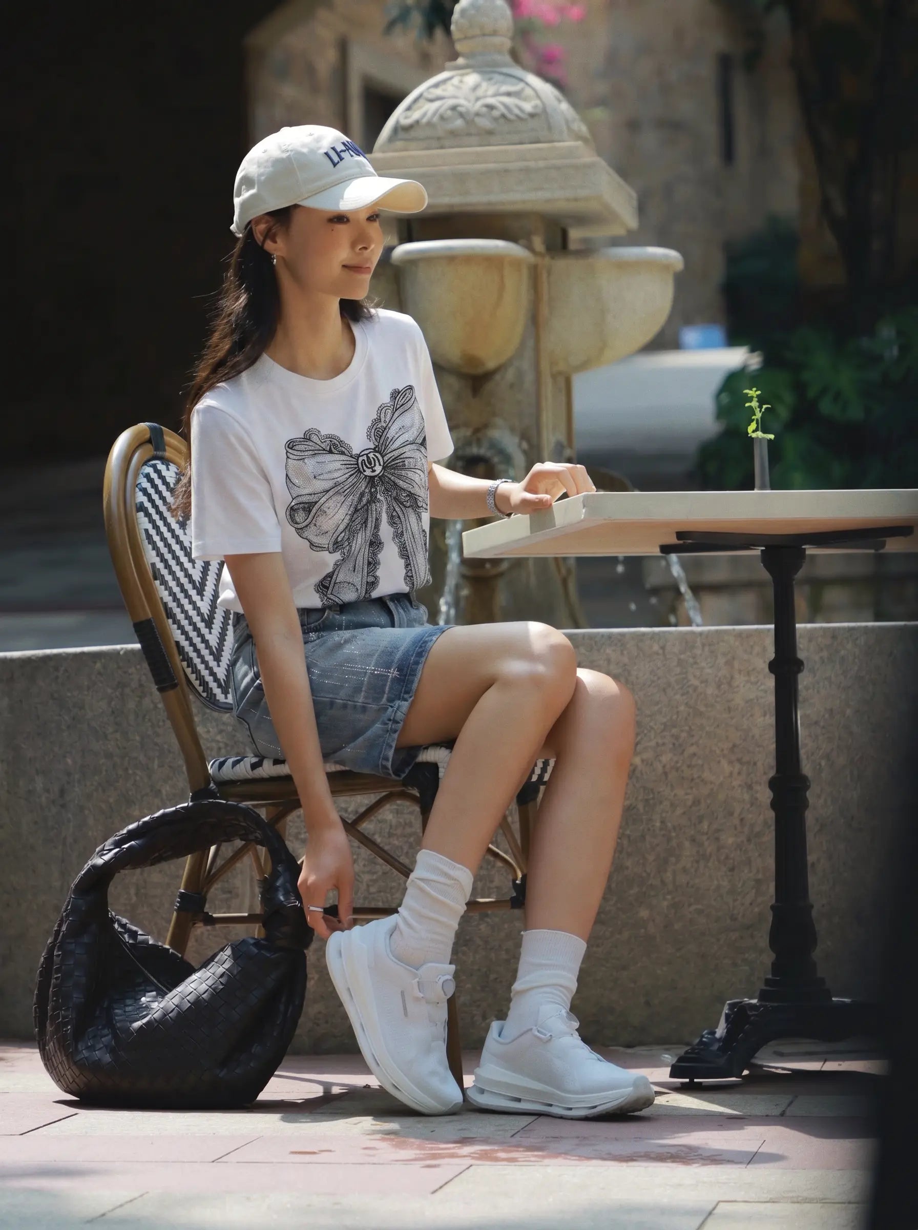 A woman sits at an outdoor cafe table wearing a white graphic t-shirt, denim shorts, white socks, and white sneakers, with a black shoulder bag beside her. TCH Rhinestone Vintage Bow Graphic White T-Shirt