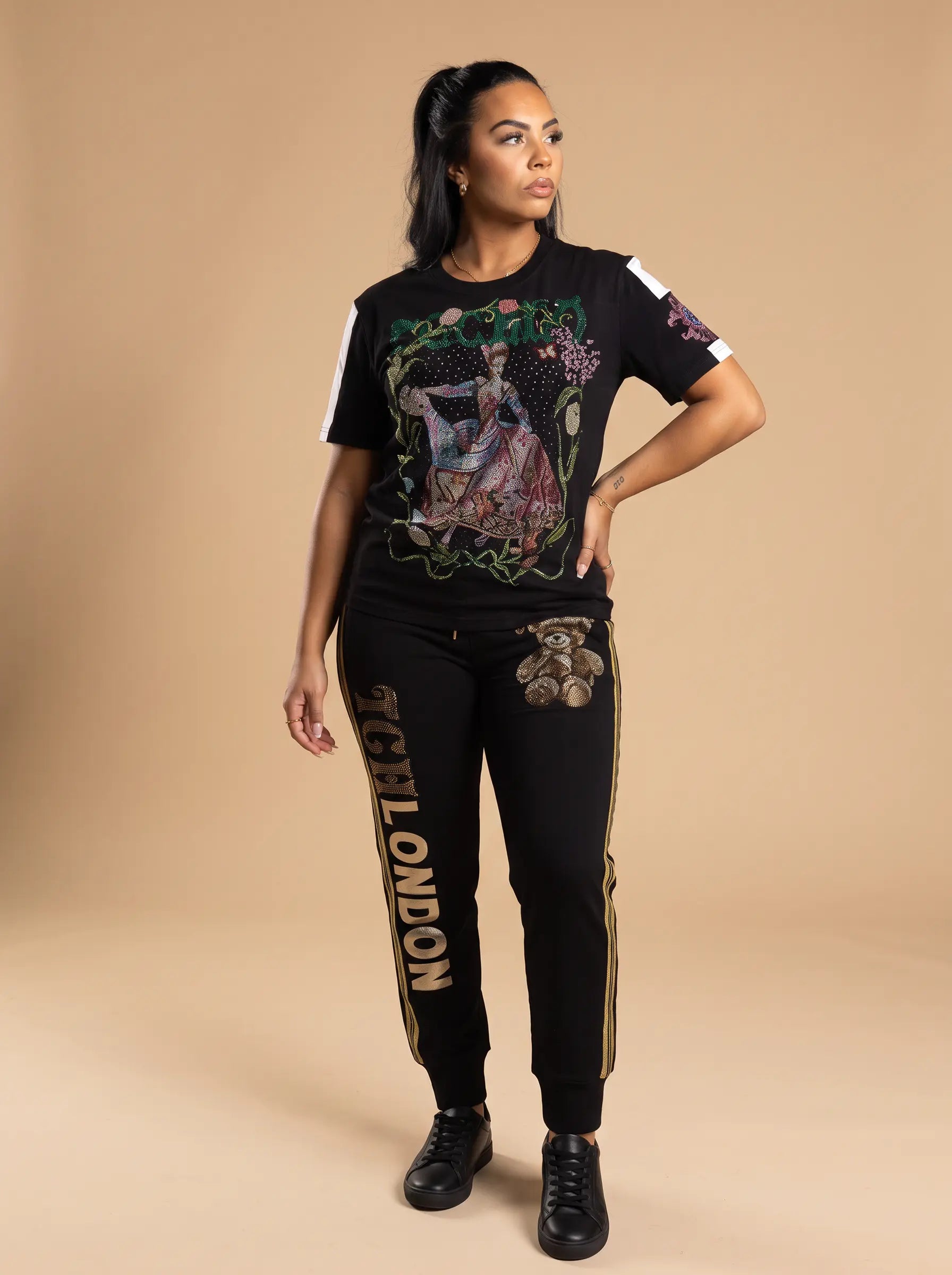 A woman modeling a black graphic t-shirt and matching black sweatpants with gold and white details, standing against a solid beige studio background. TCH Rhinestone Vintage Floral Artwork Graphic Black T-Shirt