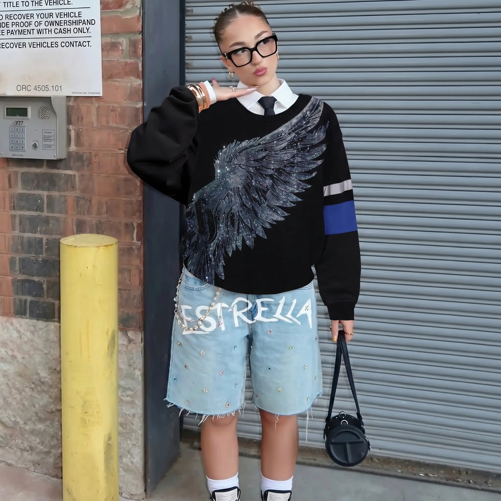 Person wearing a black sweater with a wing design, denim shorts, and holding a black bag against a garage door background. TCH Rhinestone Wing Black Crewneck Sweatshirt