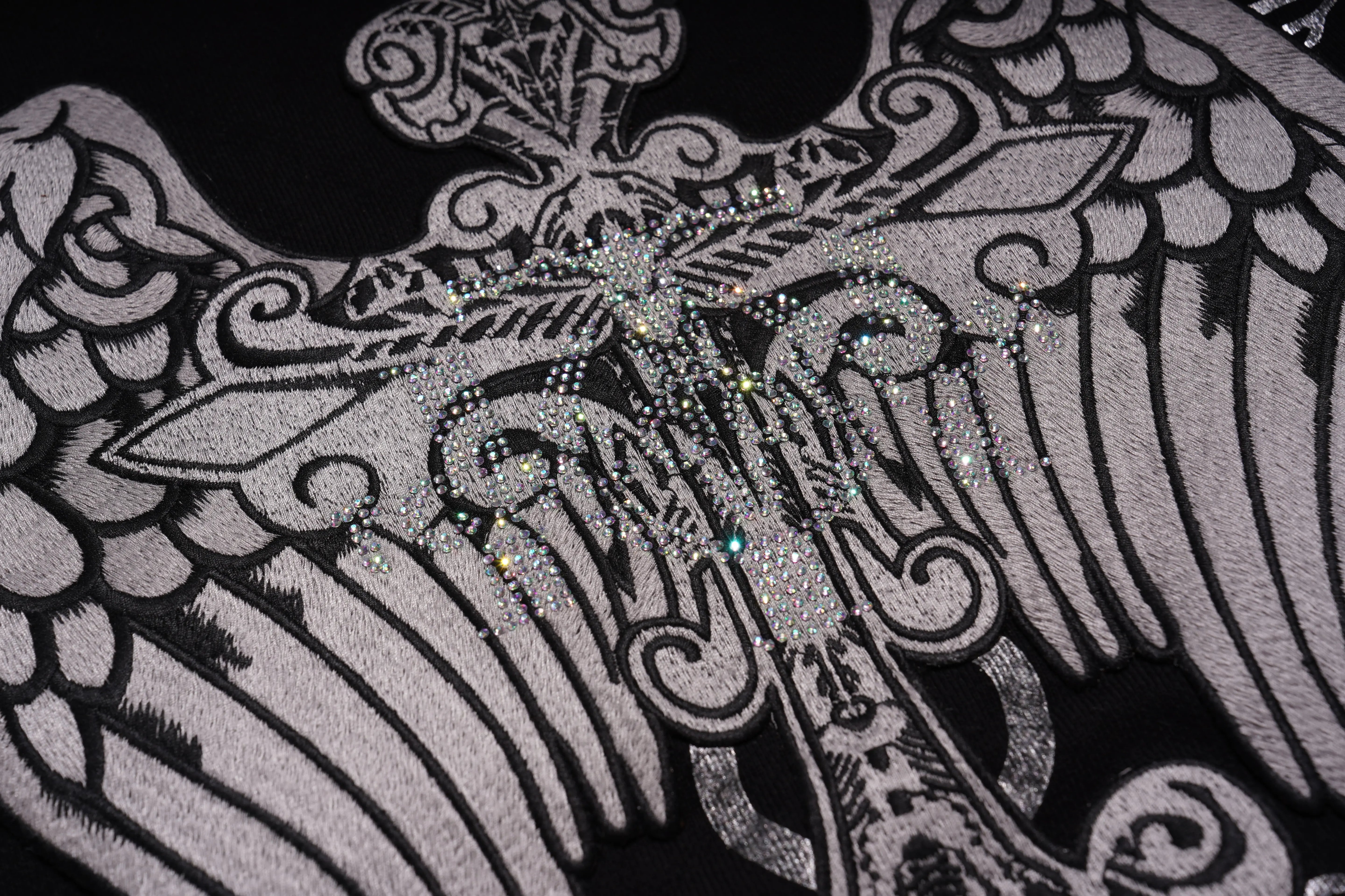 Close-up of a decorative design with intricate patterns and embellishments on a black background. TCH Rhinestone Wing Crest Embroidered Sweatshirt