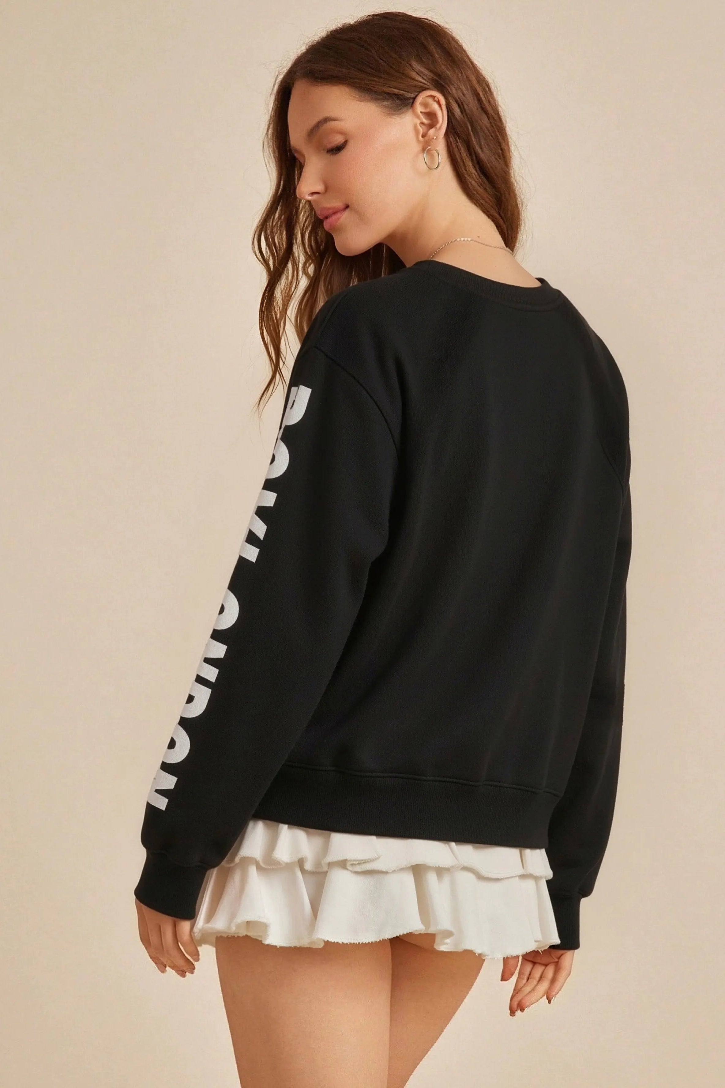 Woman wearing a black sweatshirt with white text and a white skirt on a beige background， TCH Rhinestone Wing Overlay Sweatshirt