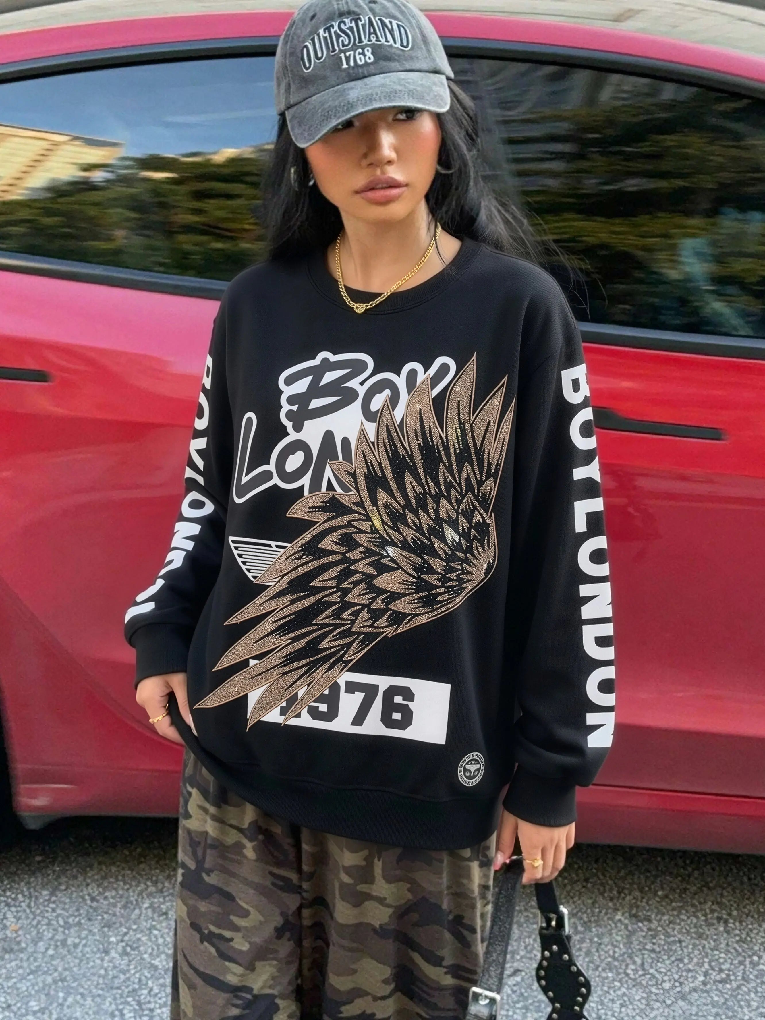 Person wearing a black sweatshirt with 'Boy London' branding and camouflage pants, standing in front of a red car. TCH Rhinestone Wing Overlay Sweatshirt