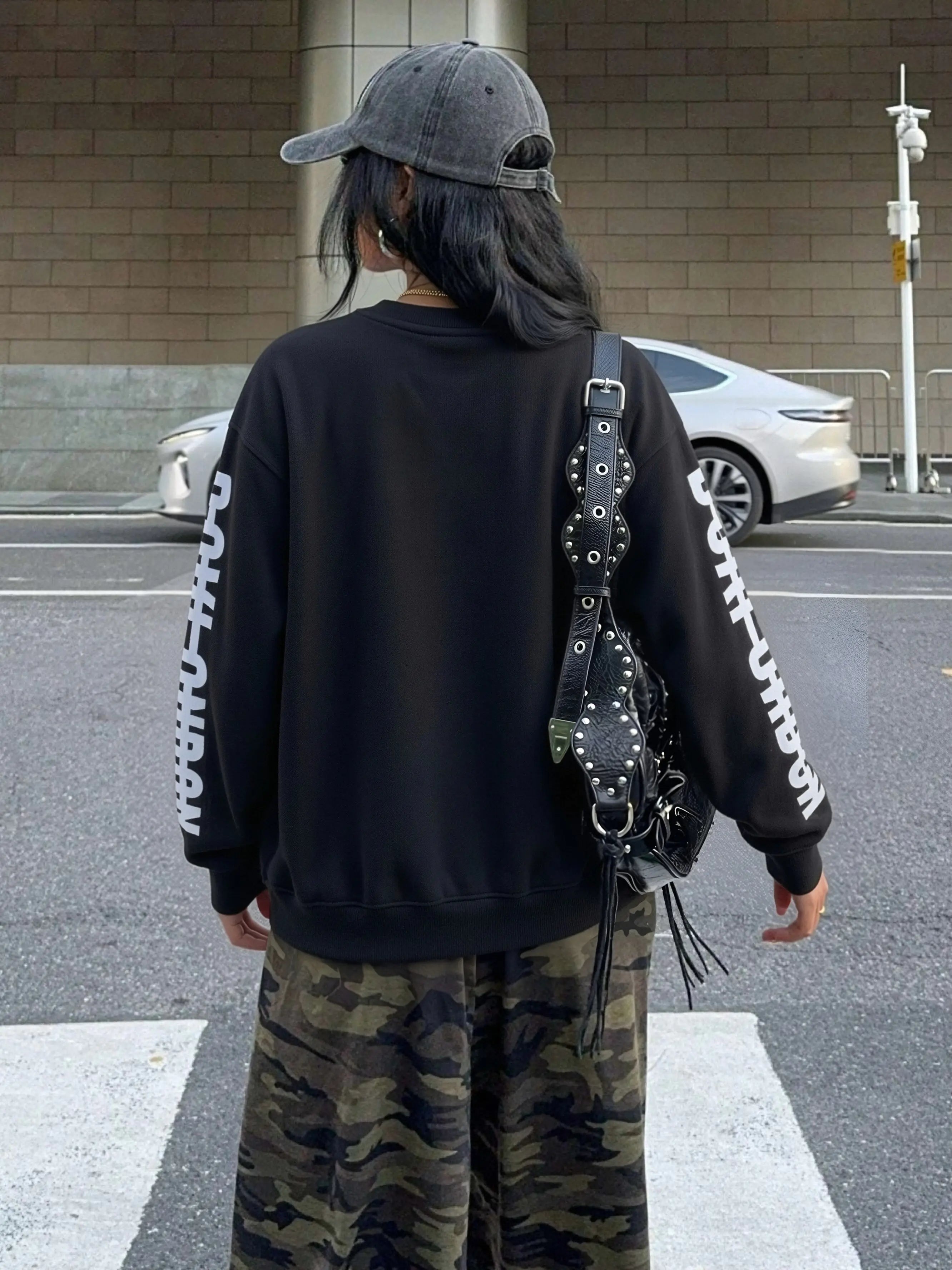 Person wearing a black sweatshirt with white text, camouflage pants, and a studded backpack on a street. TCH Rhinestone Wing Overlay Sweatshirt