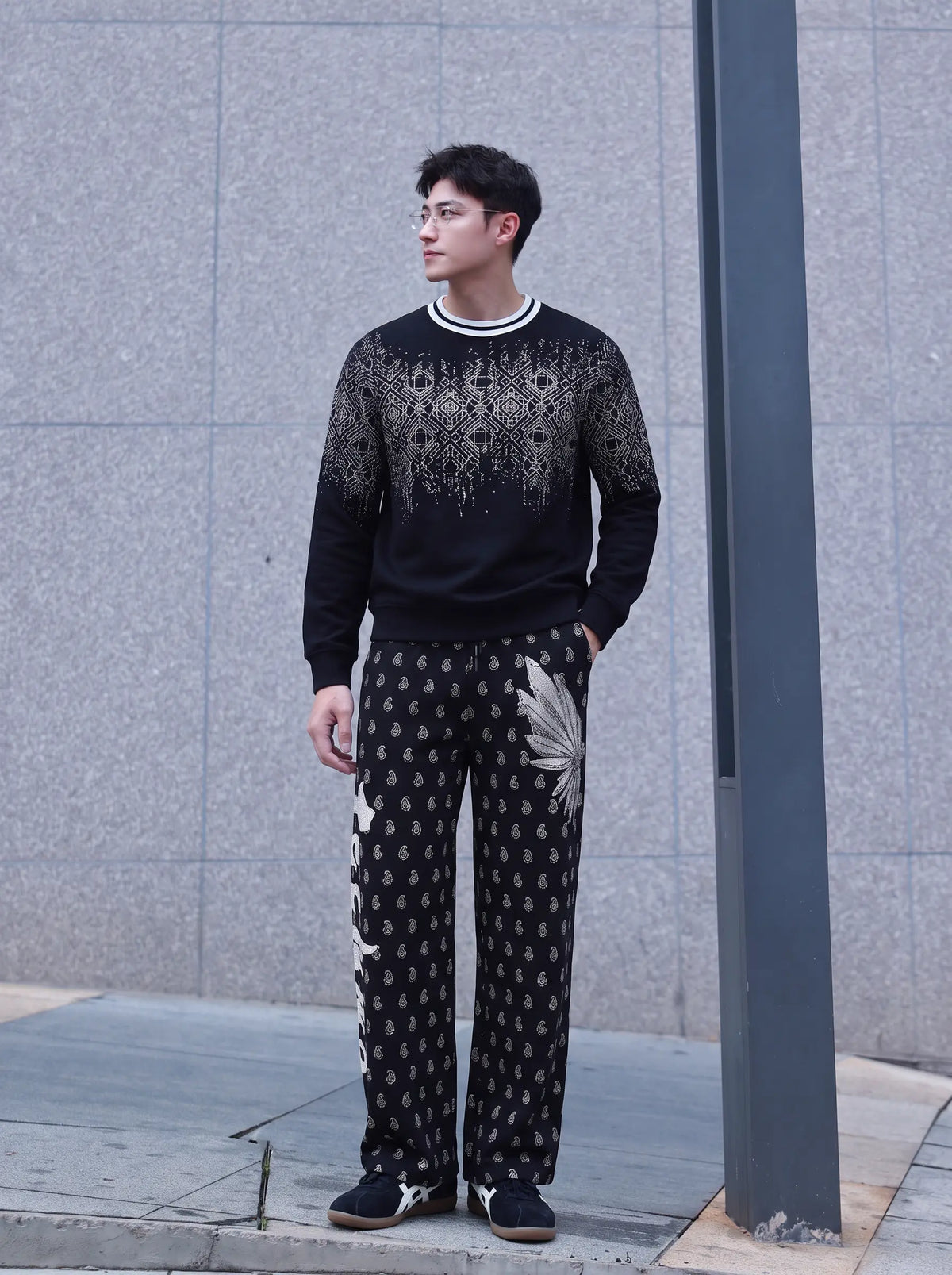 A man wearing a black crewneck sweater with a white knit pattern on the chest and black patterned trousers, standing outdoors against a gray stone wall. TCH Rhinestone Wing Paisley Wide-Leg Sweatpants