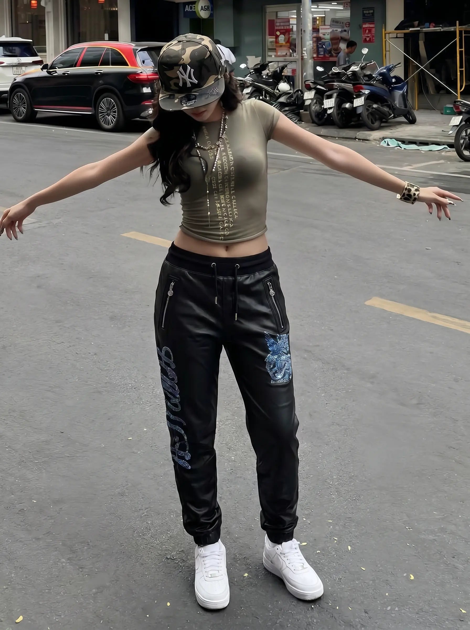 A woman stands on a city street with her arms outstretched, wearing a fitted metallic olive crop top, black distressed pants with blue graphic patches and white sneakers, and a camouflage baseball cap. TCH Rhinestone Wing Teddy Faux Leather Jogger Pants