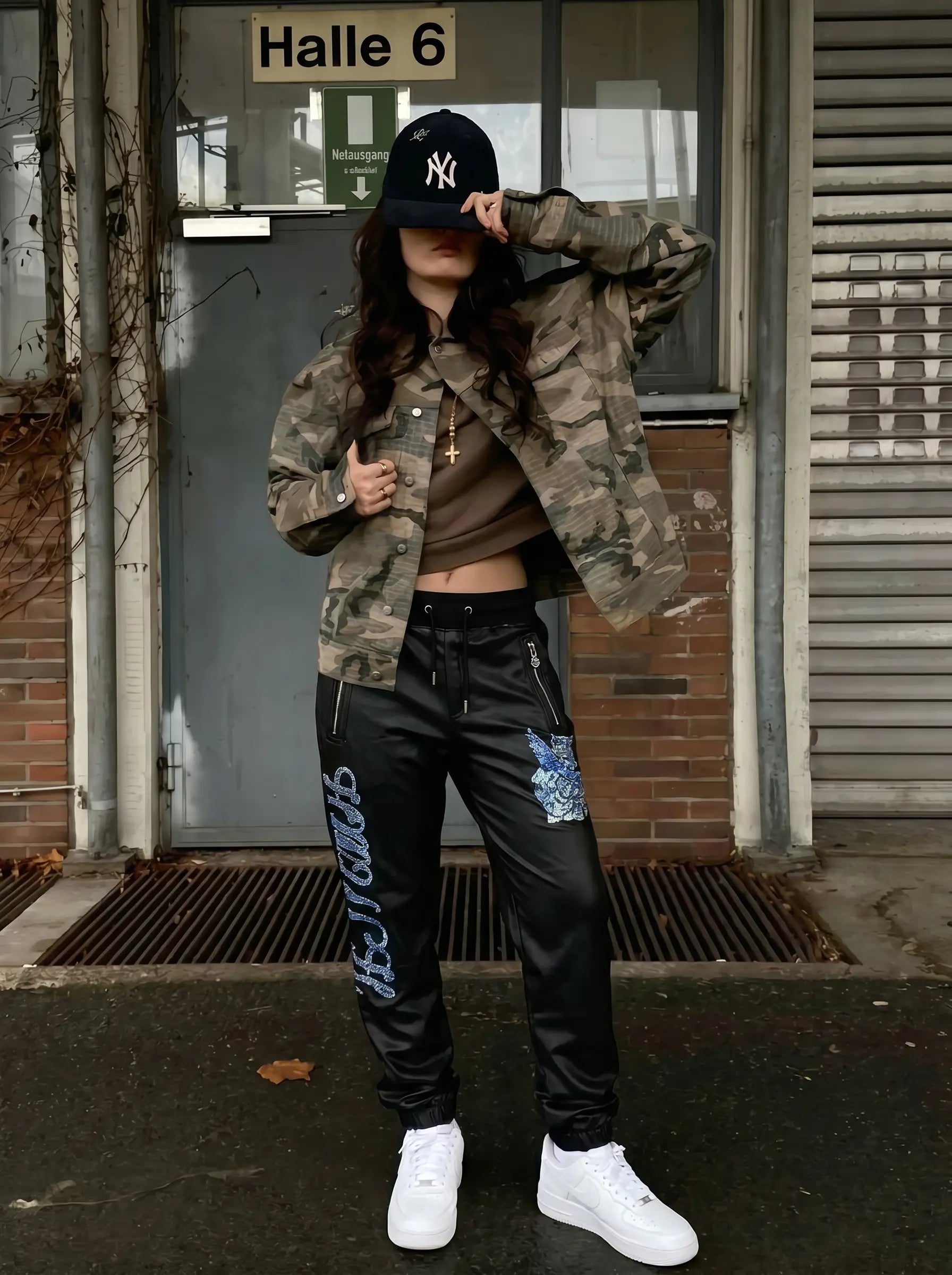 A person stands in front of a building entrance wearing a camouflage bomber jacket, black cropped pants with blue graphic details, a brown cropped top, white sneakers, and a black New York Yankees baseball cap. TCH Rhinestone Wing Teddy Faux Leather Jogger Pants