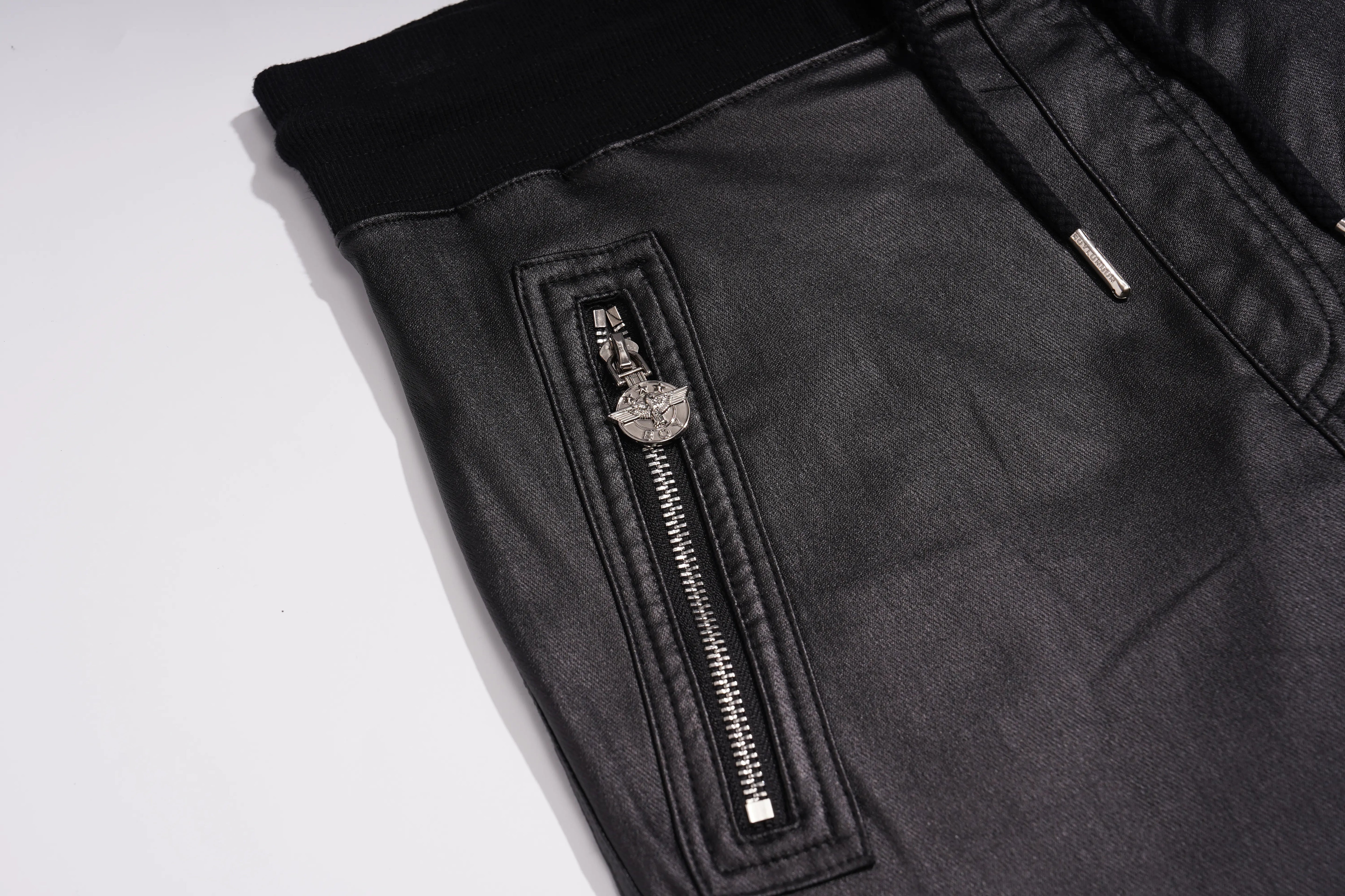 Close-up of black faux-leather pants showing a vertical zippered pocket with a silver zipper and a decorative metal pull, alongside a black elastic waistband. TCH Rhinestone Wing Teddy Faux Leather Jogger Pants
